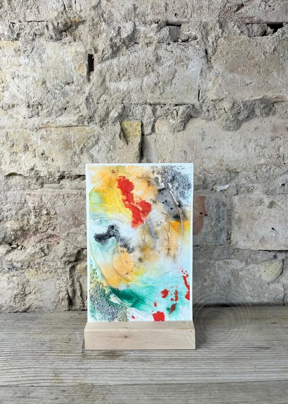 Abstract mixed media painting 'Even now I shine' in red, green, and gold, displayed on wooden stand against rustic wall.