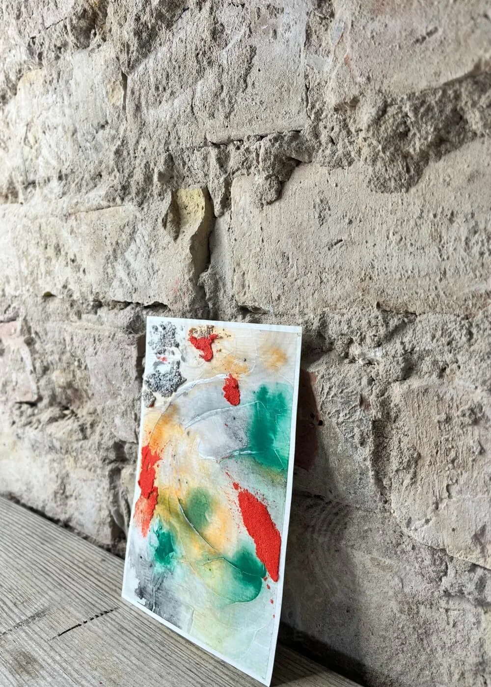 Angled view of abstract artwork leaning against a rough stone wall, vibrant colors standing out against the neutral background.