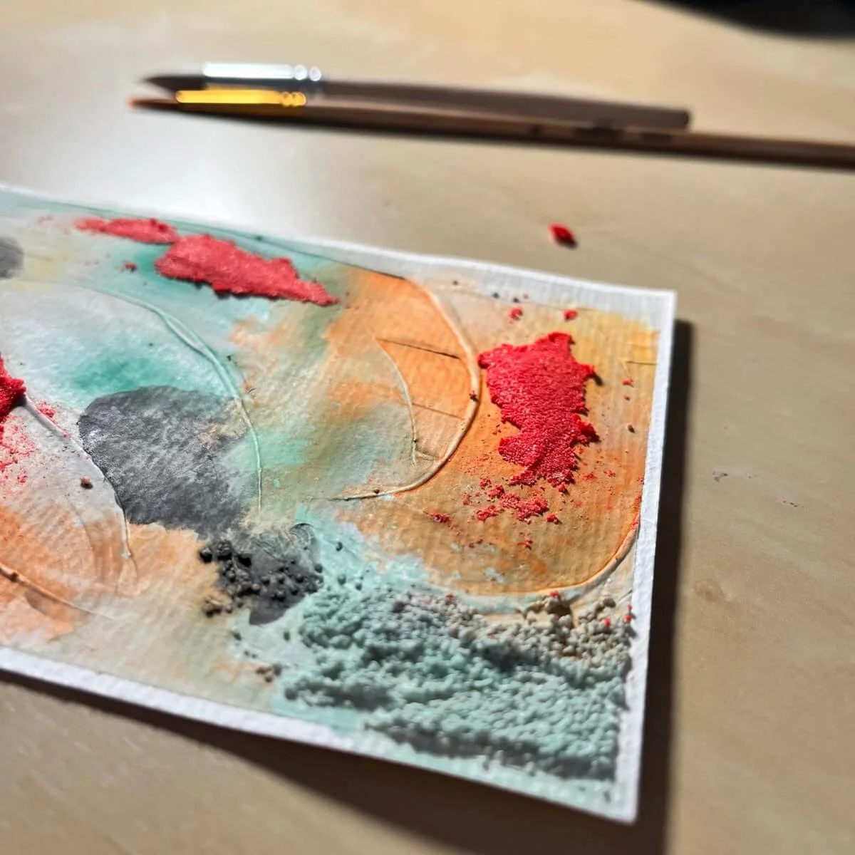 Close-up of a textured abstract artwork from the Built Of Light series before the silver finish was applied. The painting features vivid red, orange, green, and charcoal pigments. Two fine brushes lie in the soft-focus background