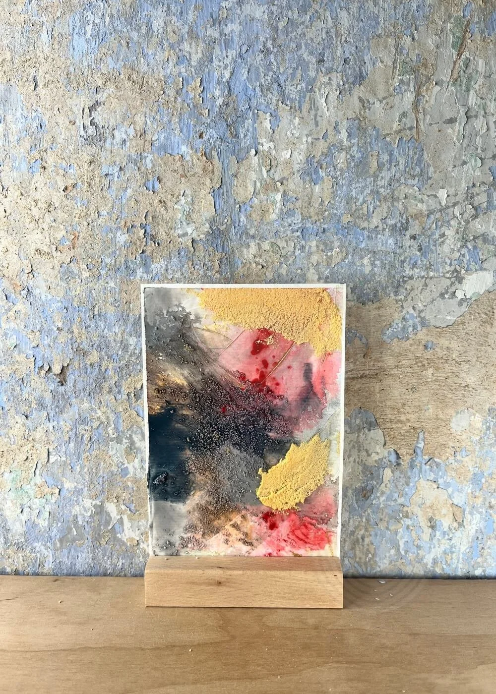 An abstract artwork stands against a softly worn wall — gold and crimson layers melt into smoke-grey stillness, embodying quiet feminine strength.