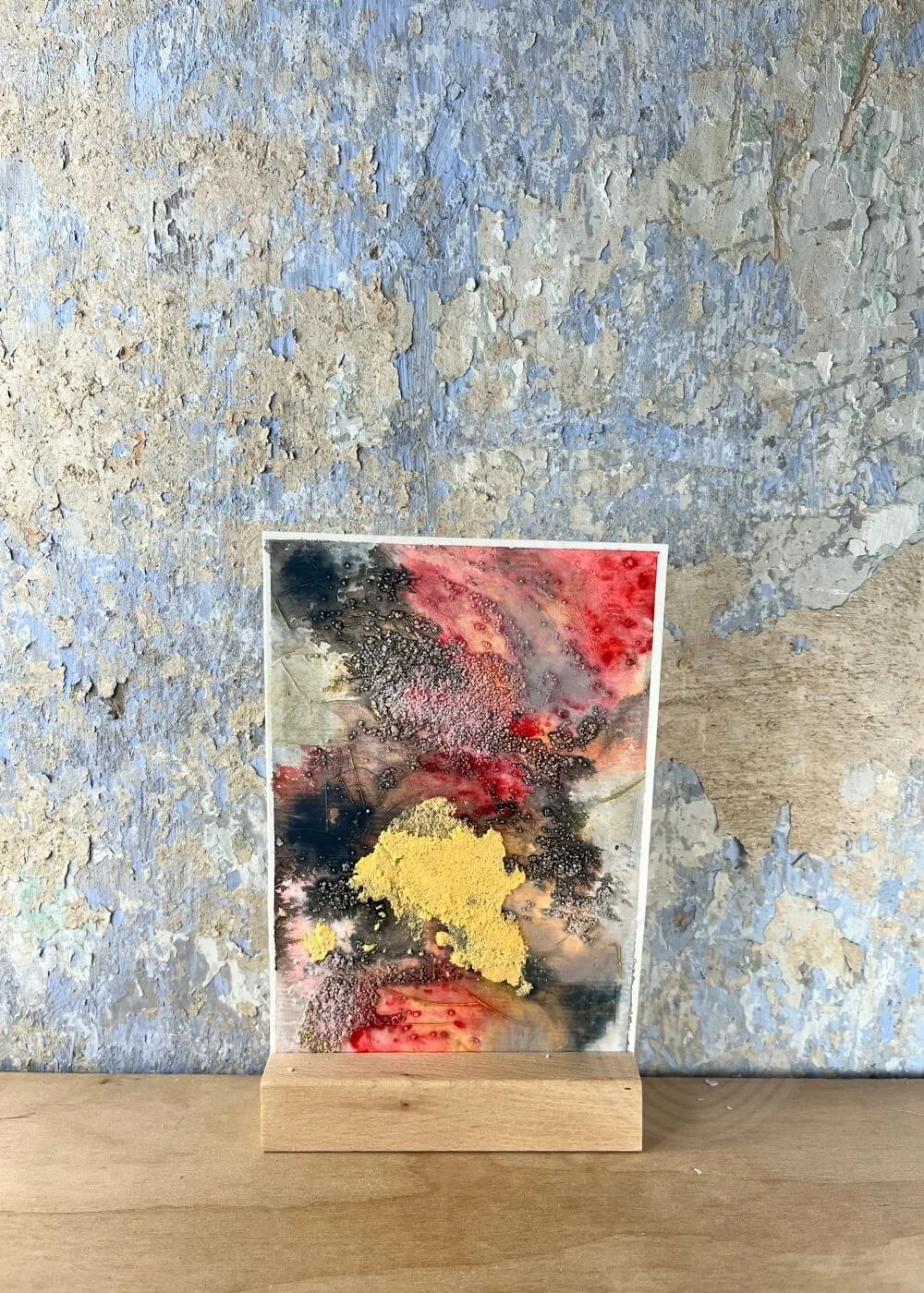 A luminous mixed-media artwork stands against a weathered blue wall — a quiet storm of crimson, gold, and charcoal whispering of what once stayed unspoken.