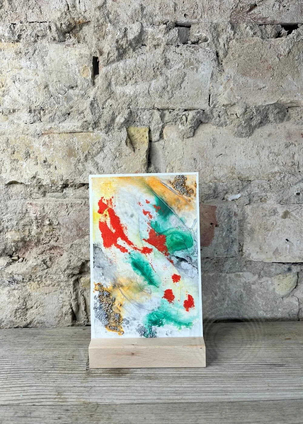 Abstract mixed media artwork "Where I Draw the Line" with textured red, green, gold, and black pigments on canvas board, displayed on wooden stand against stone wall.