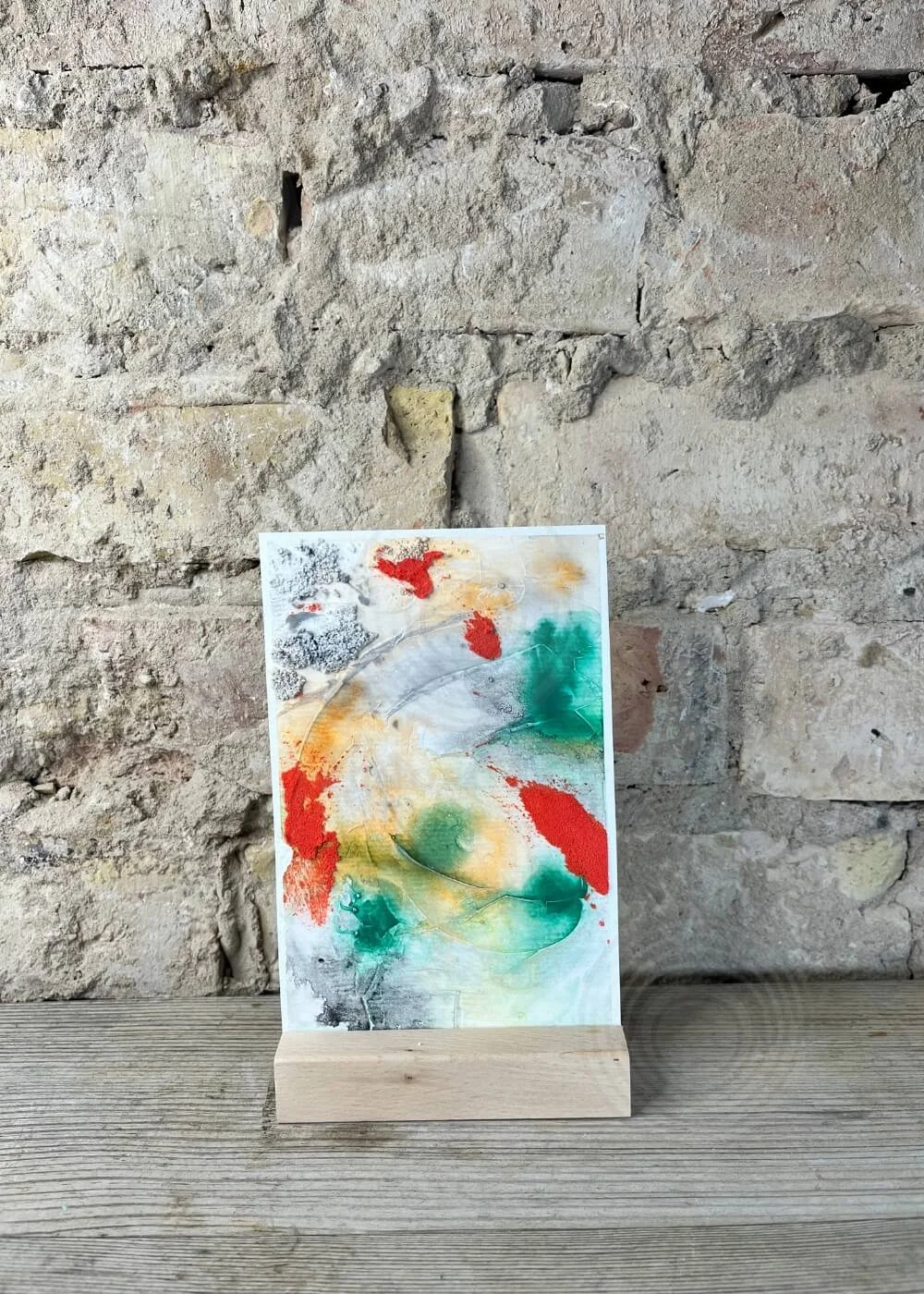 Abstract mixed-media artwork in a wooden stand against a natural stone wall, featuring vivid red, green, and orange tones contrasted with grey textures.