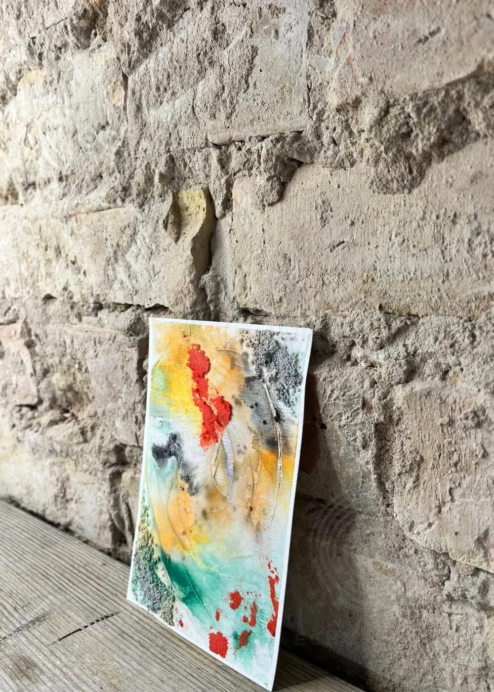 Side view of textured abstract artwork Even Now I Shine, leaning against stone wall with vivid colors.