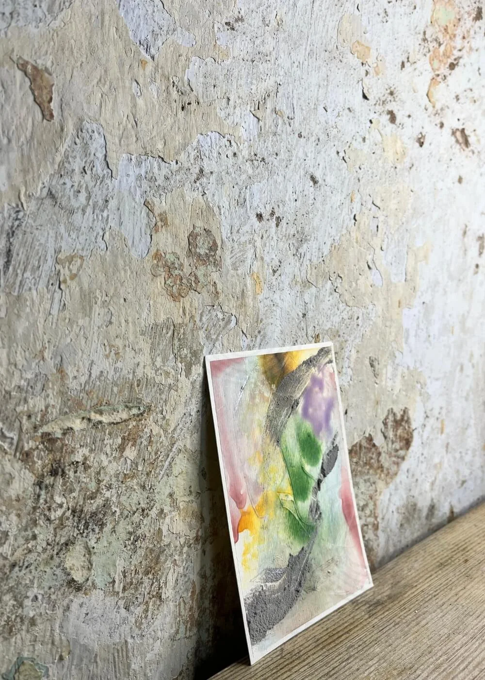 Abstract artwork "Between the Seconds No. 8" with layered textures in green, yellow, purple, and grey, shown leaning slightly against a rough, weathered wall.
