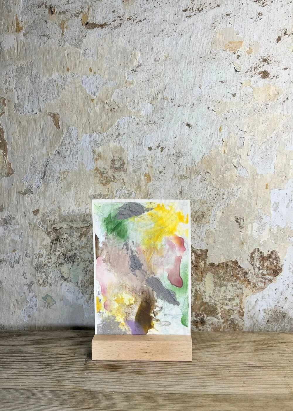 Abstract artwork "Between the Seconds No. 6" on a small canvas with soft green, yellow, pink, and textured grey tones, standing in a wooden holder against a rustic wall.