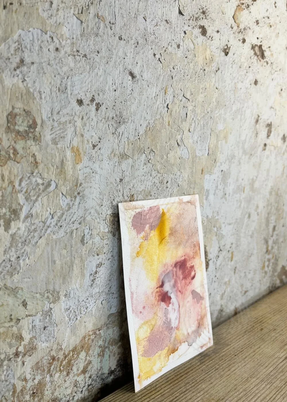 Abstract artwork “Held by Myself No. 8” in rose, burgundy, and golden yellow tones, shown from a side angle leaning against a rustic wall.