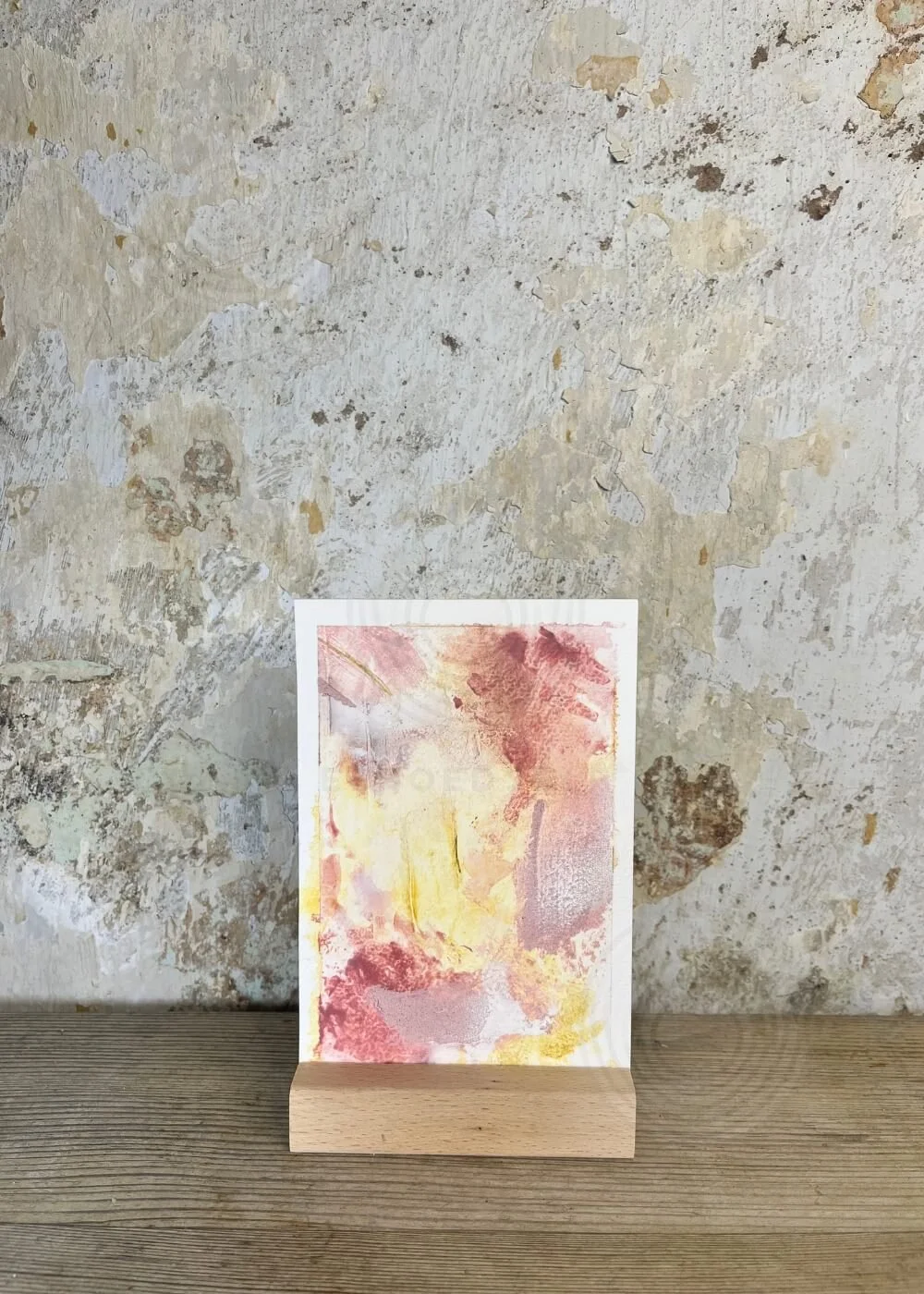 Abstract art card "Held by myself No. 9" in soft rosé and golden yellow tones, displayed on a wooden stand against a textured wall.