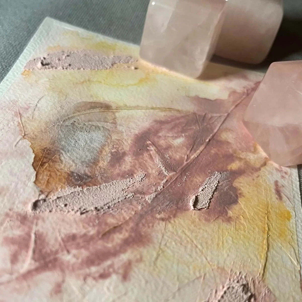 Close-up of textured mixed-media artwork in soft pink and yellow hues, paired with three raw rose quartz crystals – symbolic mood image from the Held By Myself series