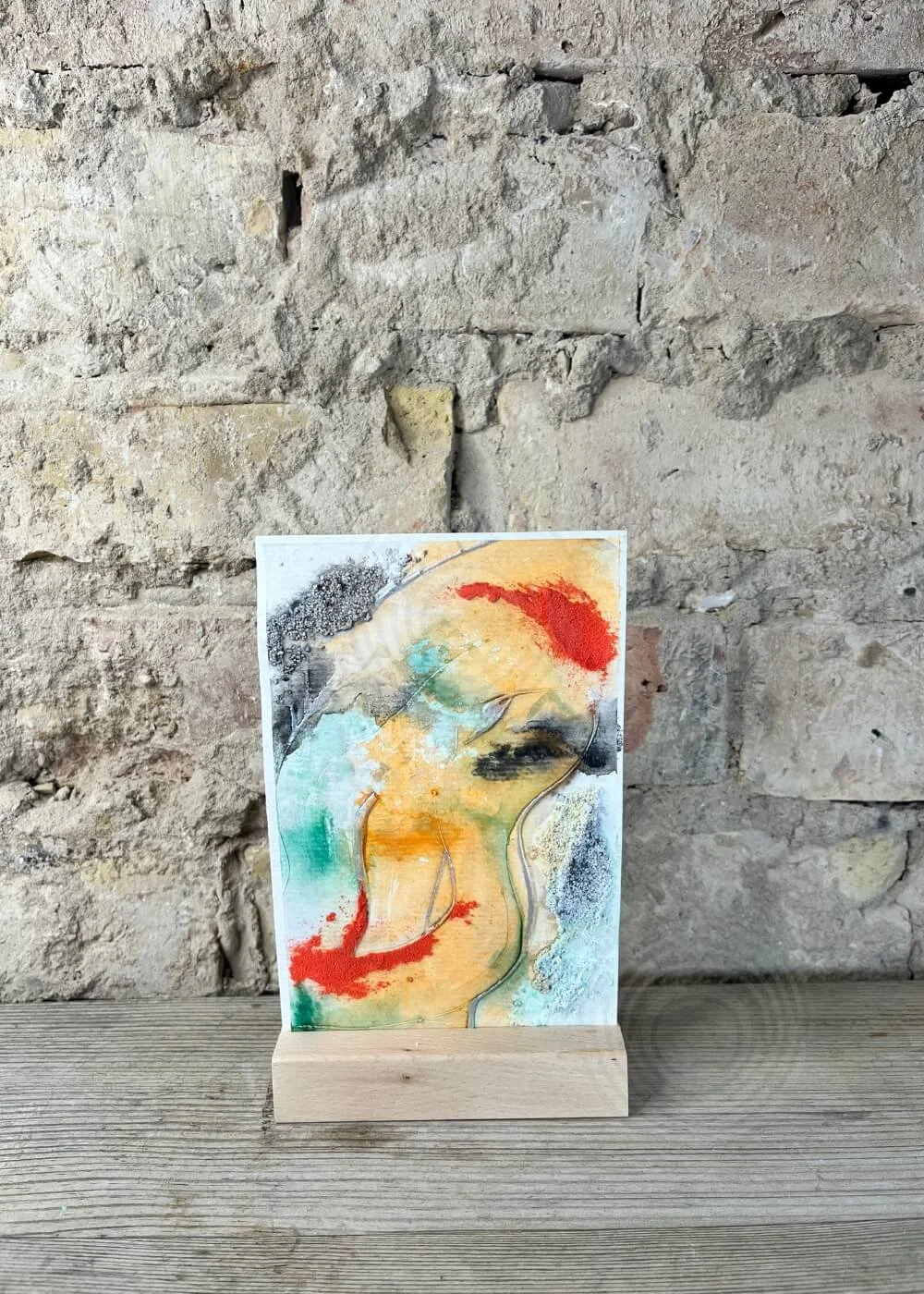 Abstract artwork Where I end and begin from the Built of Light series, textured layers in orange, green, black and red pigments against a rustic stone wall, displayed in a wooden stand.