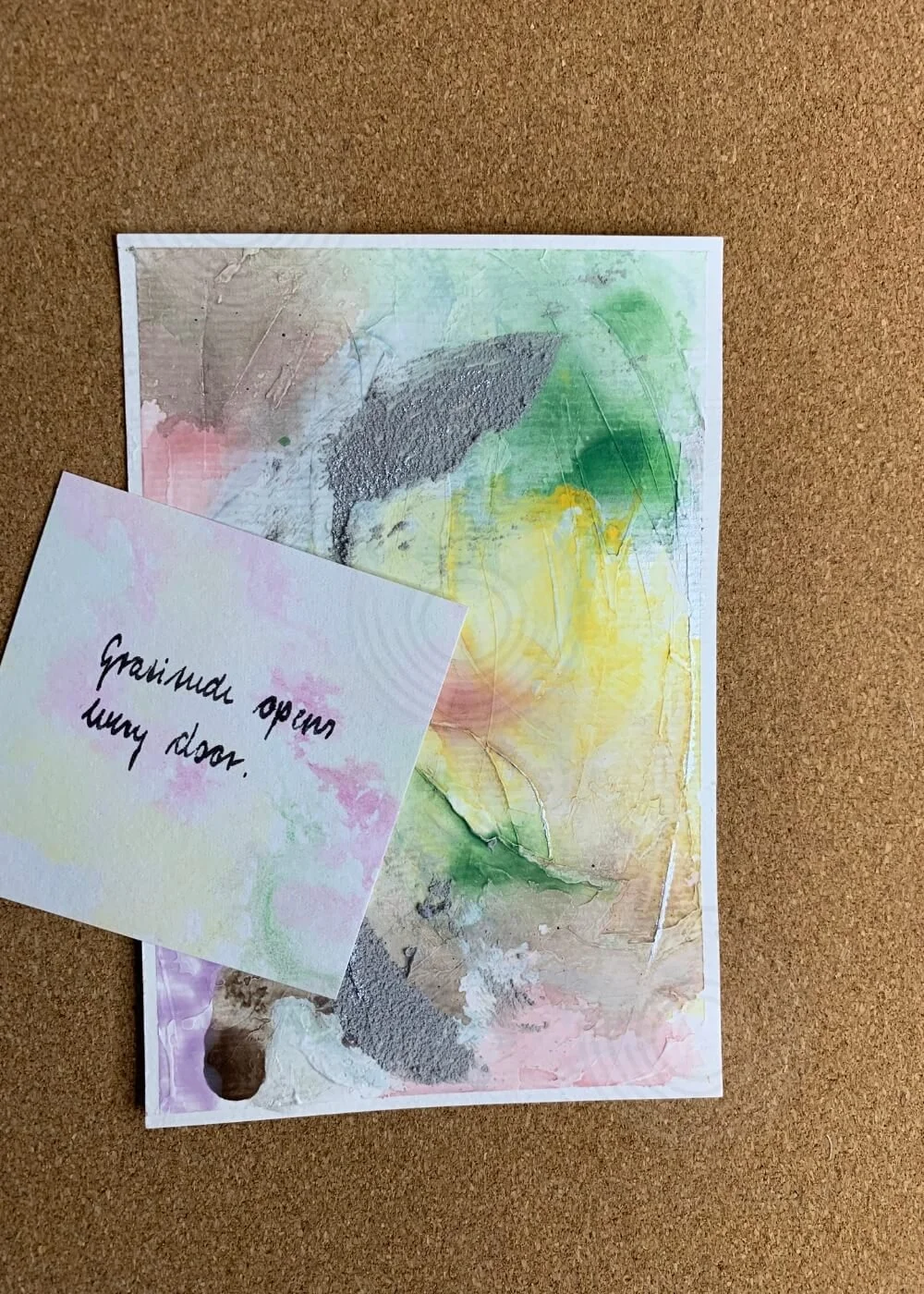 Abstract mixed-media artwork "Between the Seconds No. 7" in pastel tones with affirmation card reading ‘Gratitude opens every door’ on cork background.