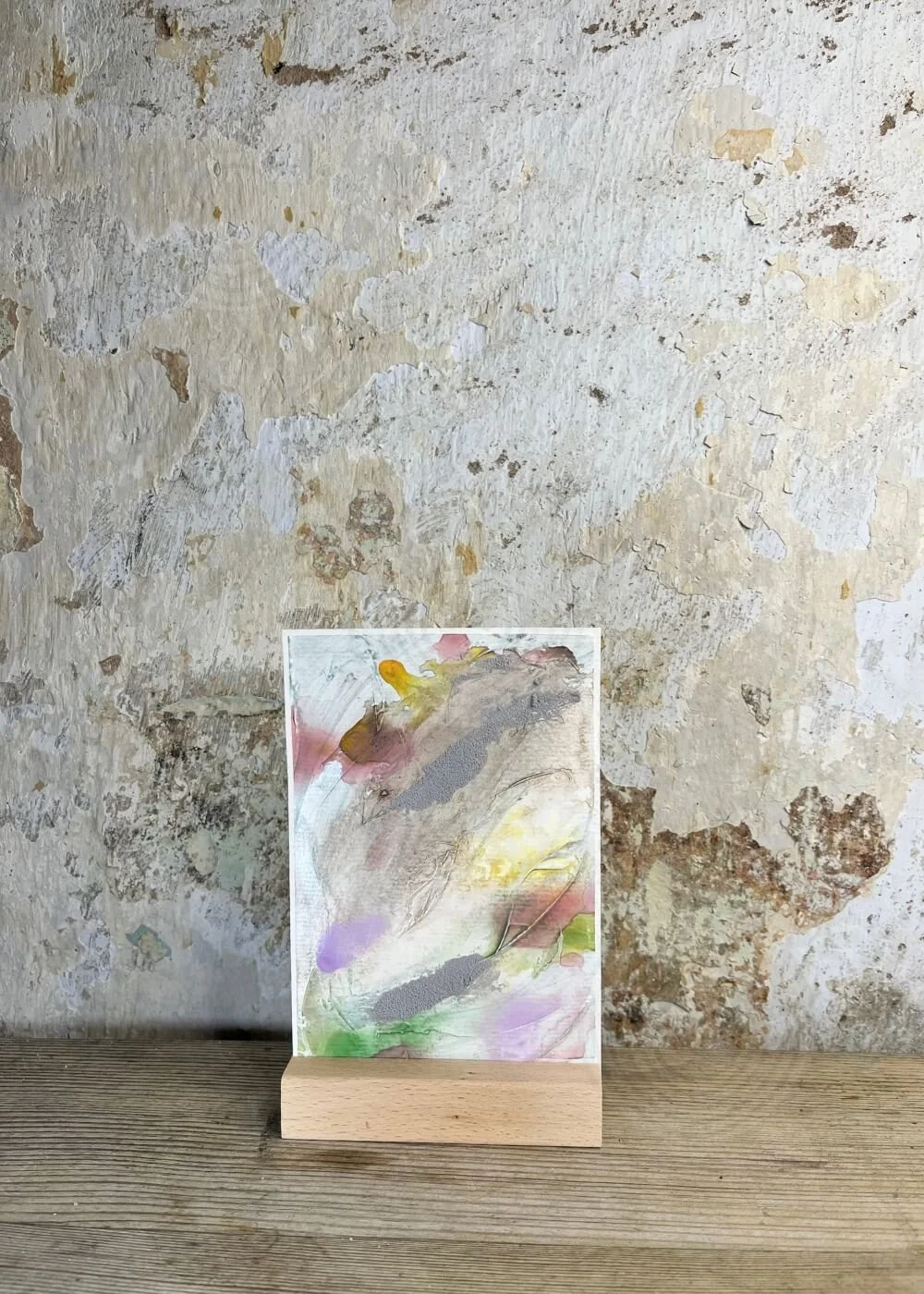 Front view of “Between the Seconds No. 5,” abstract mixed-media artwork in beige with lavender, green and yellow accents, textured surface on paper, displayed on a wooden stand.