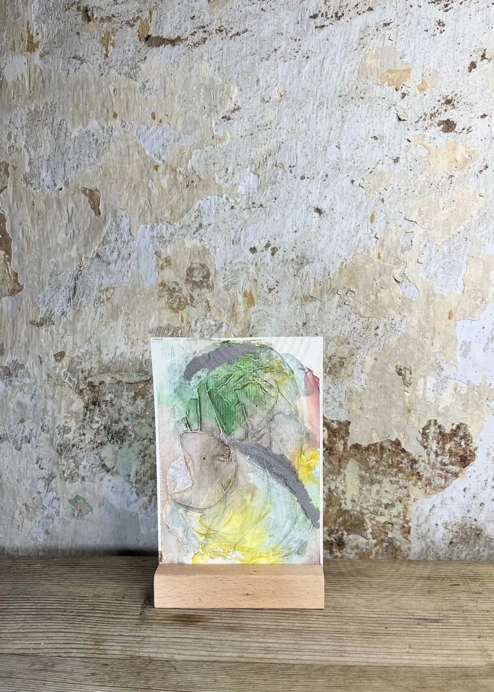 Abstract mixed-media artwork "Between the Seconds No. 10"  in soft green, yellow, grey and rose tones, displayed upright on a wooden stand against a textured rustic wall.