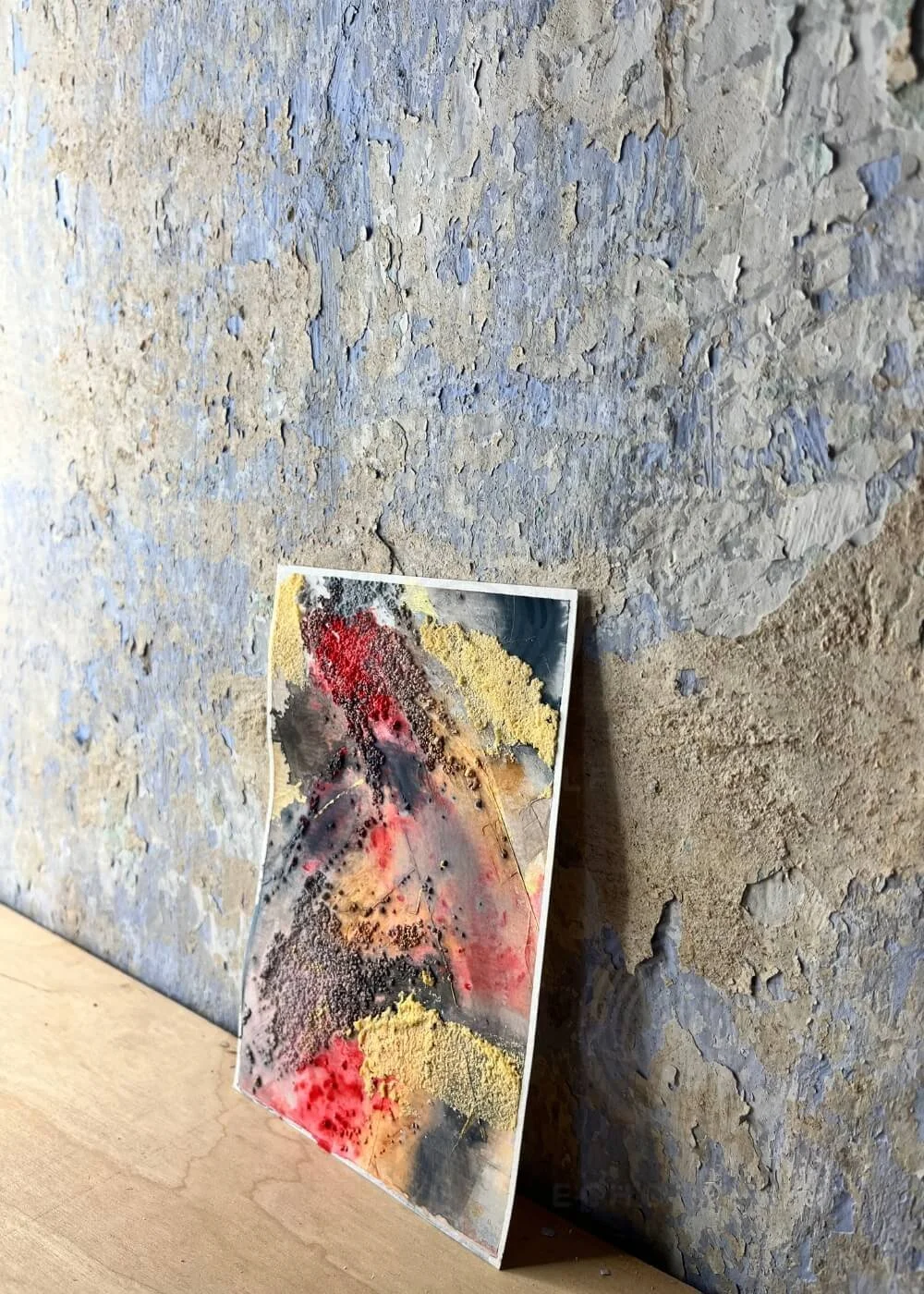 The painting leans against a weathered wall, its edges breathing quiet resilience — a reminder that healing often begins beneath what once burned.