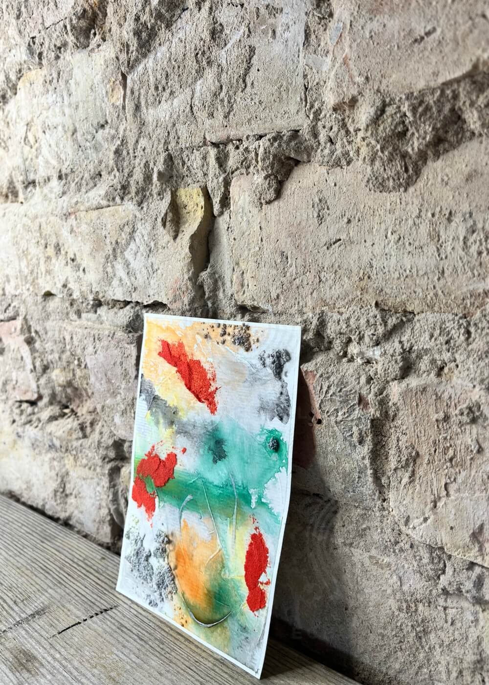 Side view of abstract mixed media painting "The light I let in" – layers of green, orange, and red with textured depth against a rustic stone wall.