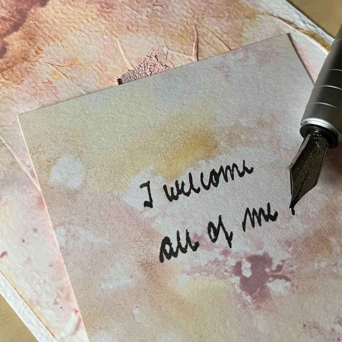 Close-up of a handwritten affirmation card that reads 'I welcome all of me' placed on textured artwork in warm pastel tones, with a fountain pen – mood image from the Held By Myself series