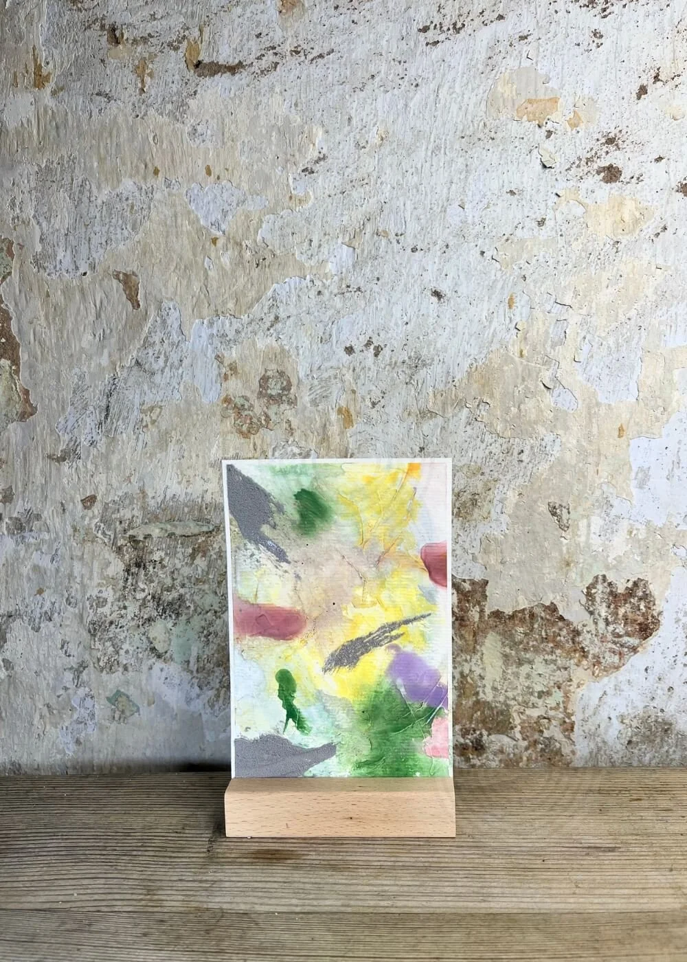 Front view of an abstract mixed-media artwork "Between the Seconds No. 3"  in green, yellow, lilac, and grey tones, displayed upright on a wooden stand against a textured wall.