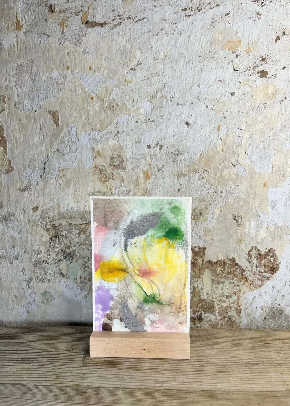 Abstract mixed-media artwork "Between the Seconds No. 7"  in pastel tones with yellow, green, and rose accents, displayed on a wooden stand against a textured wall.