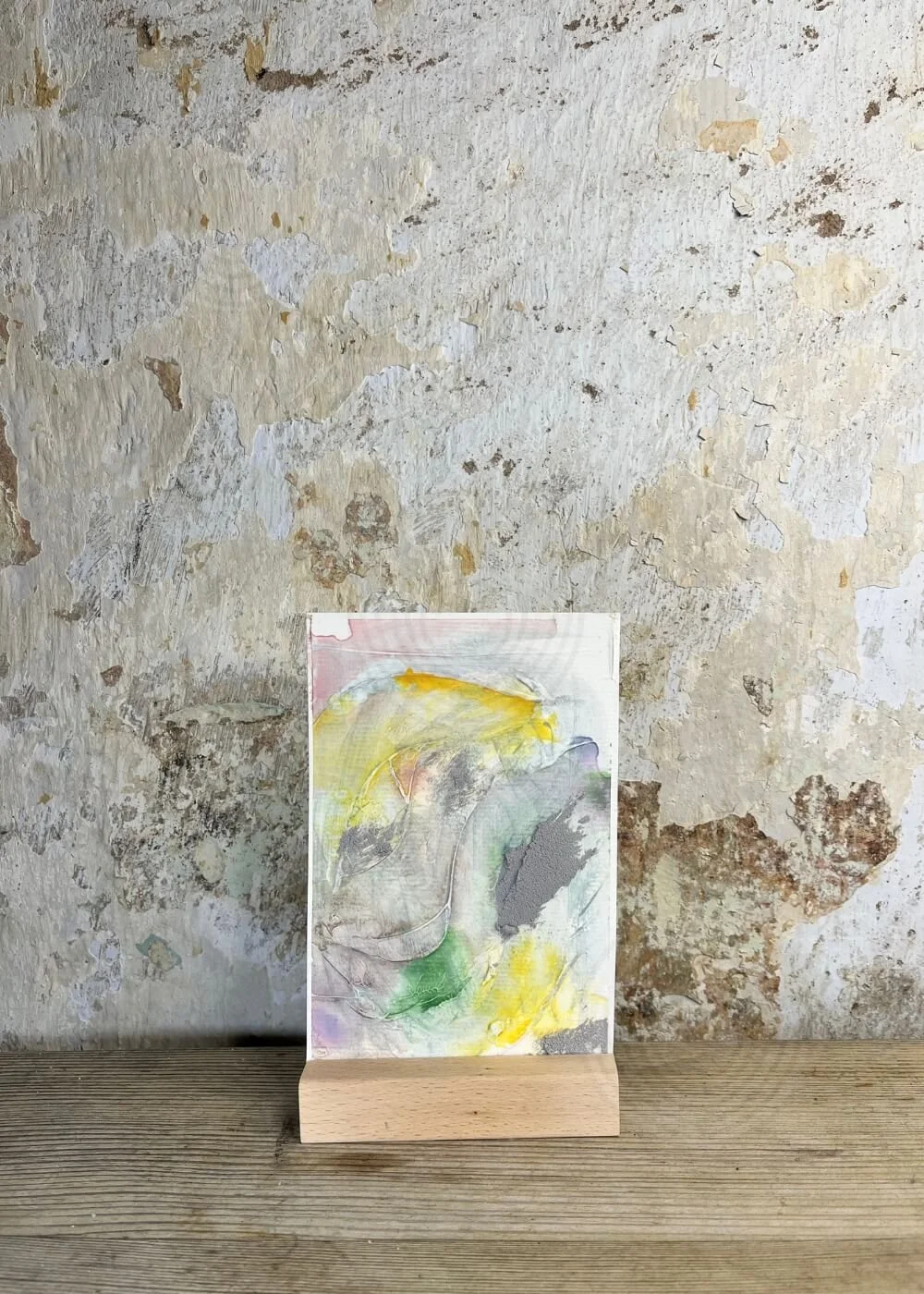 Small-format abstract artwork "Held by Myself No. 4" with soft yellow, green, and grey layers, displayed on a wooden stand against a rustic wall.