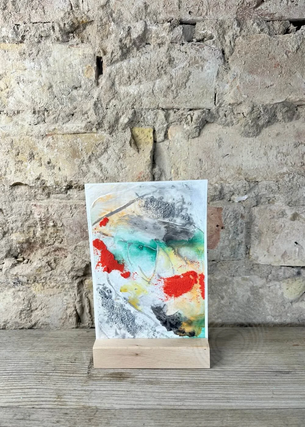 Abstract artwork “Soft shields & silent strength” displayed on wooden stand, textured background wall, colors of red, green, yellow, black and grey.