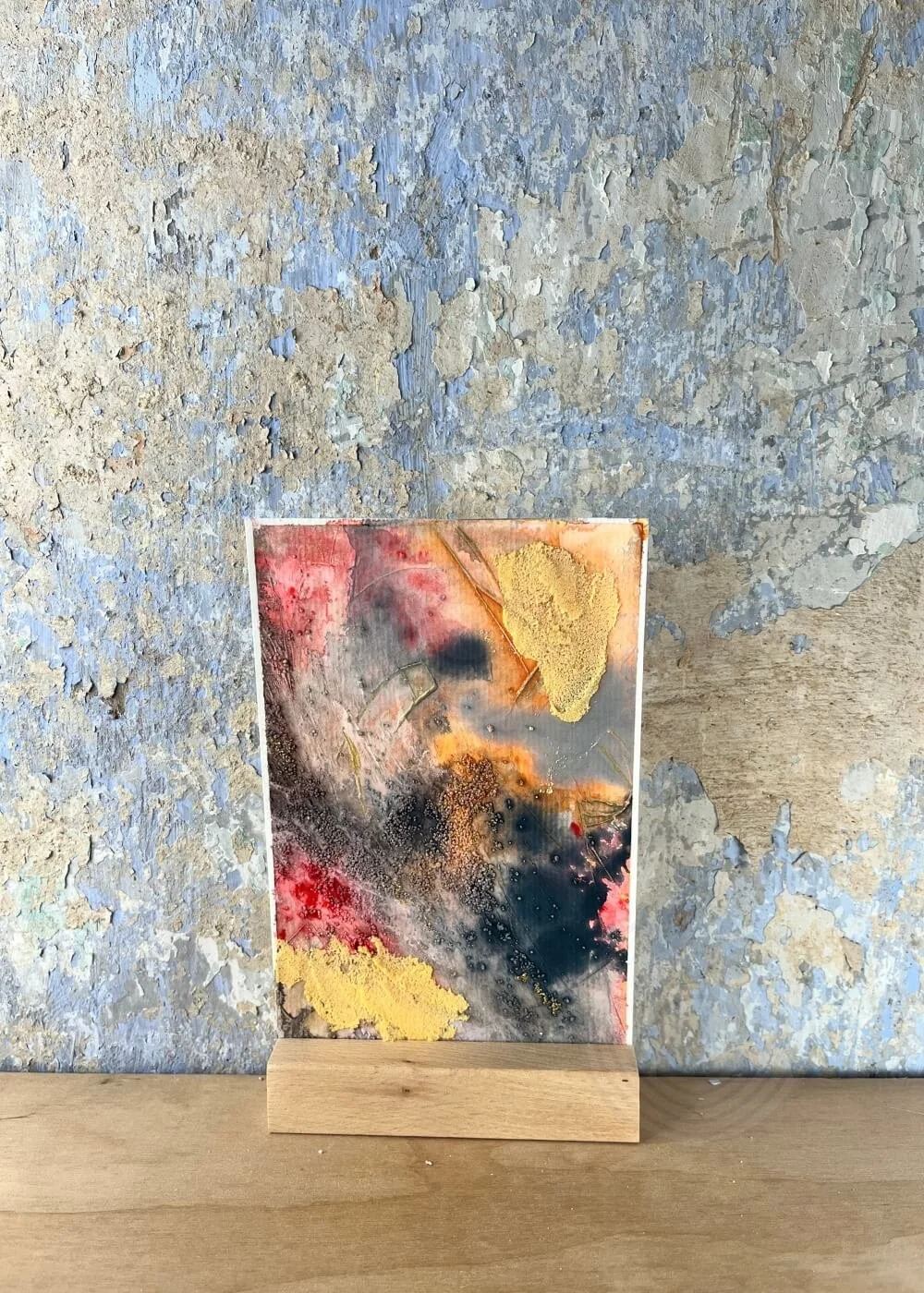 An abstract artwork rests softly against a weathered wall — traces of red, gold, and charcoal rise like smoke learning how to breathe again.