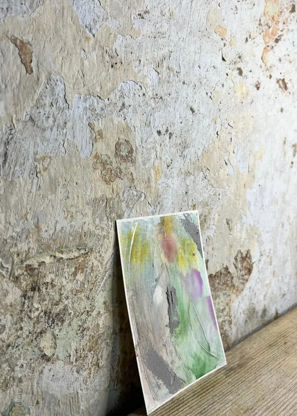 Side angle of an abstract mixed-media artwork "Between the Seconds No. 2"  with layered textures in green, purple, and yellow, leaning against a rustic wall.