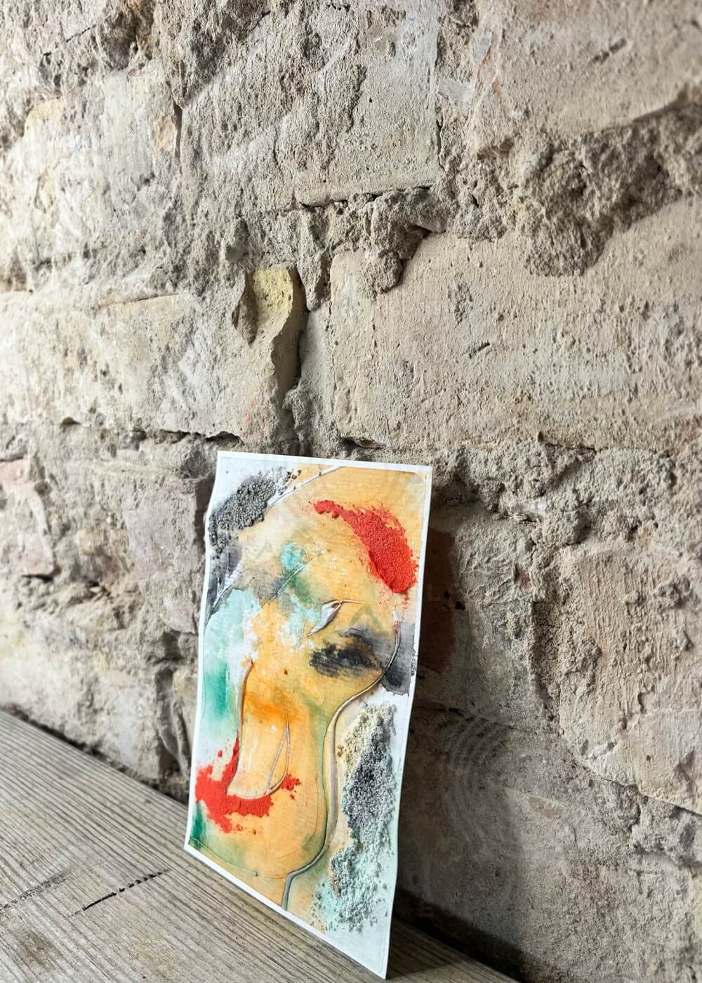 Side view of abstract painting Where I end and begin, green and orange hues with bold red details and gray textures, leaning against a stone wall.