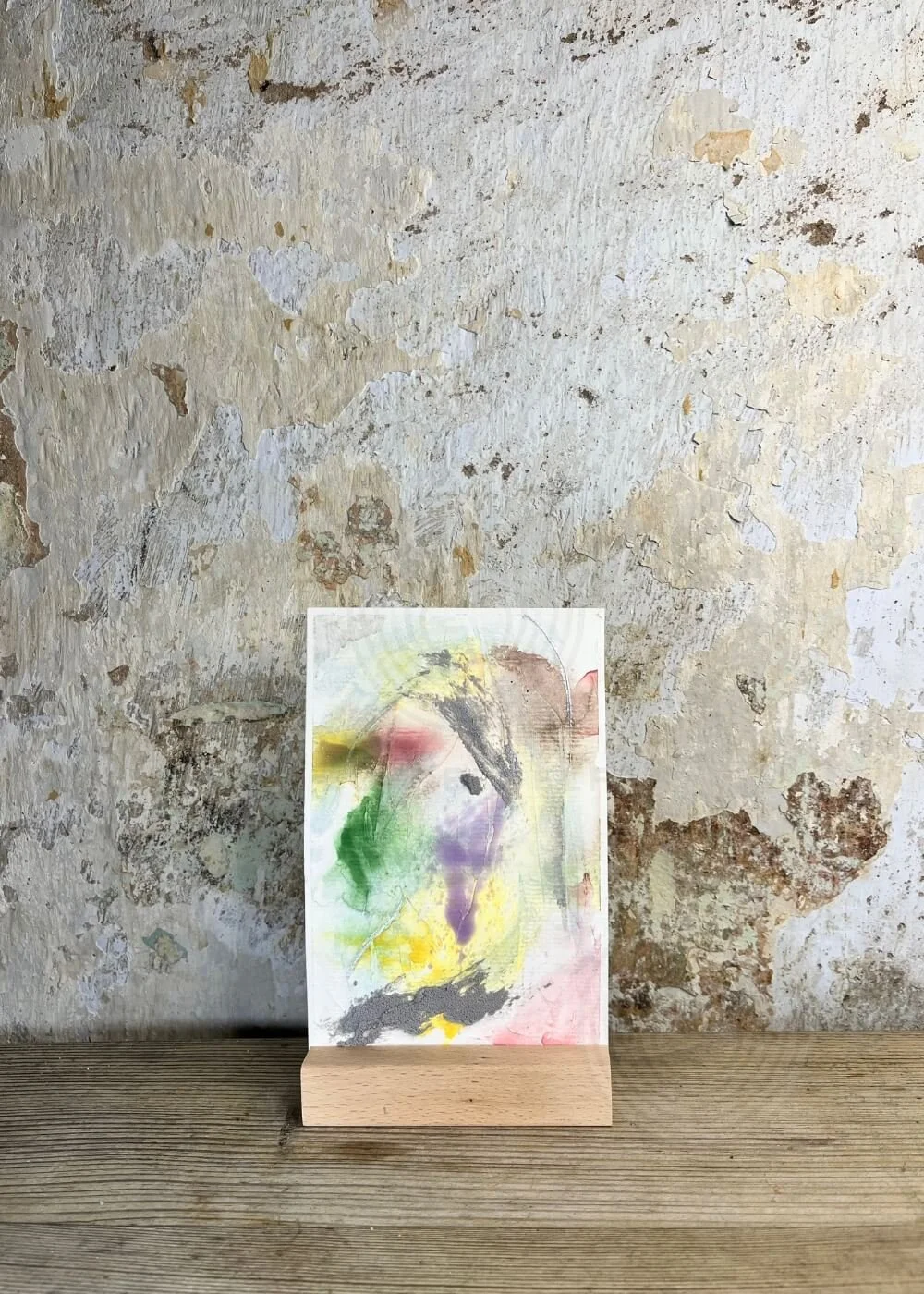 Abstract mixed media artwork No. 1 in green, yellow, purple and soft grey tones, placed on a wooden stand against a textured wall background.