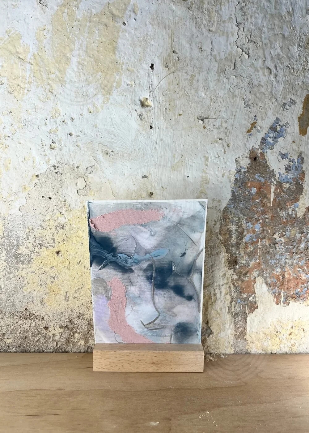 Abstract mixed-media artwork titled Between What Once Was in soft pink, grey, and blue tones — displayed against a rustic wall, radiating serenity and emotional depth.