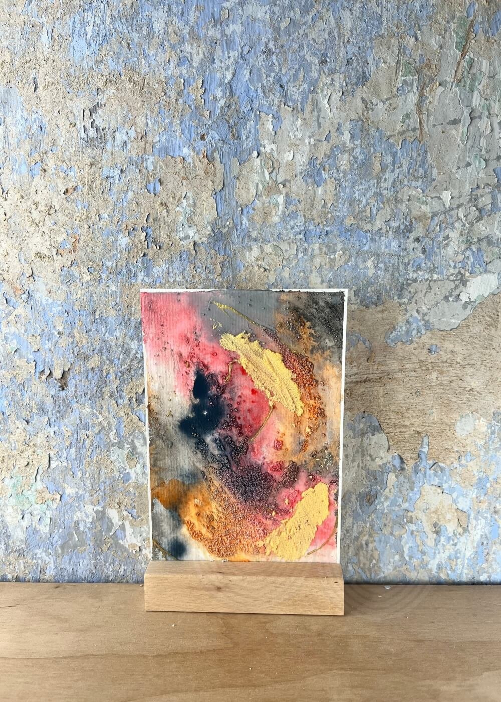 A radiant abstract artwork stands softly against a weathered wall — hues of rose, gold, and charcoal swirl like quiet flames remembering how to dance.