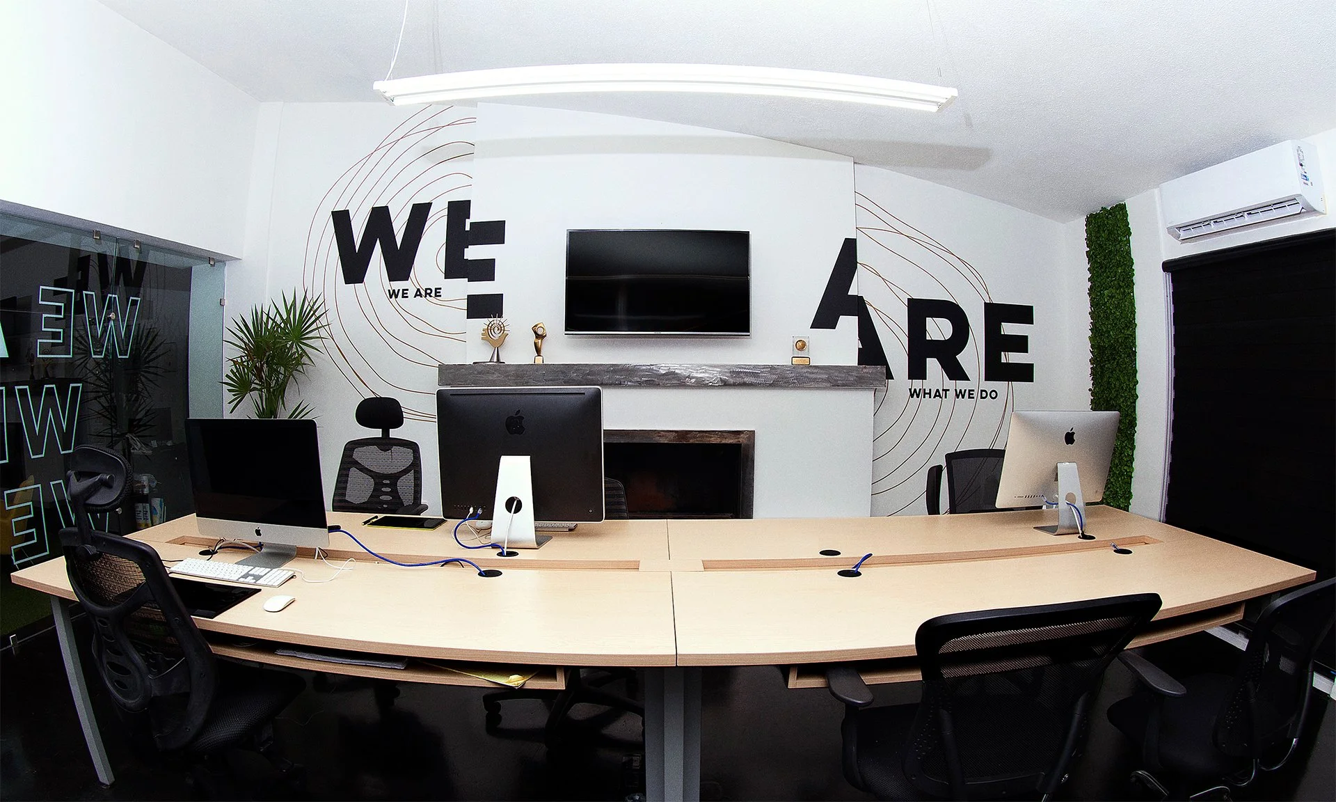 Sala de reuniones moderna con paredes blancas y decoración inspiradora que dice "We Are" y "What We Do", con mesas de madera, computadoras Apple, sillas negras, plantas y un televisor en la pared.