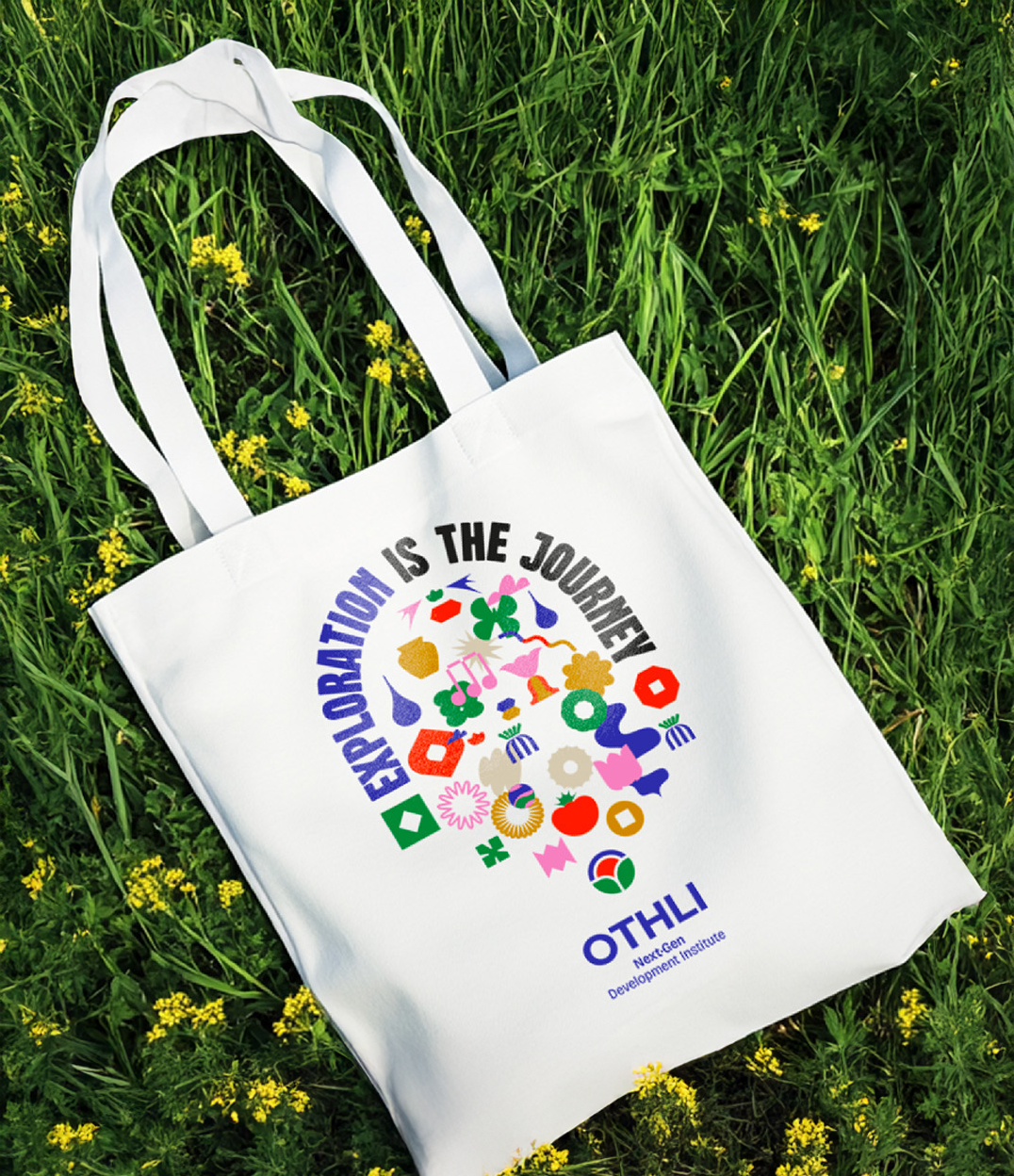 Una bolsa de tela blanca con un diseño colorido y el texto 'Exploration is the Journey' y 'OTHLI NextGen Development Institute' sobre césped verde con pequeñas flores amarillas.