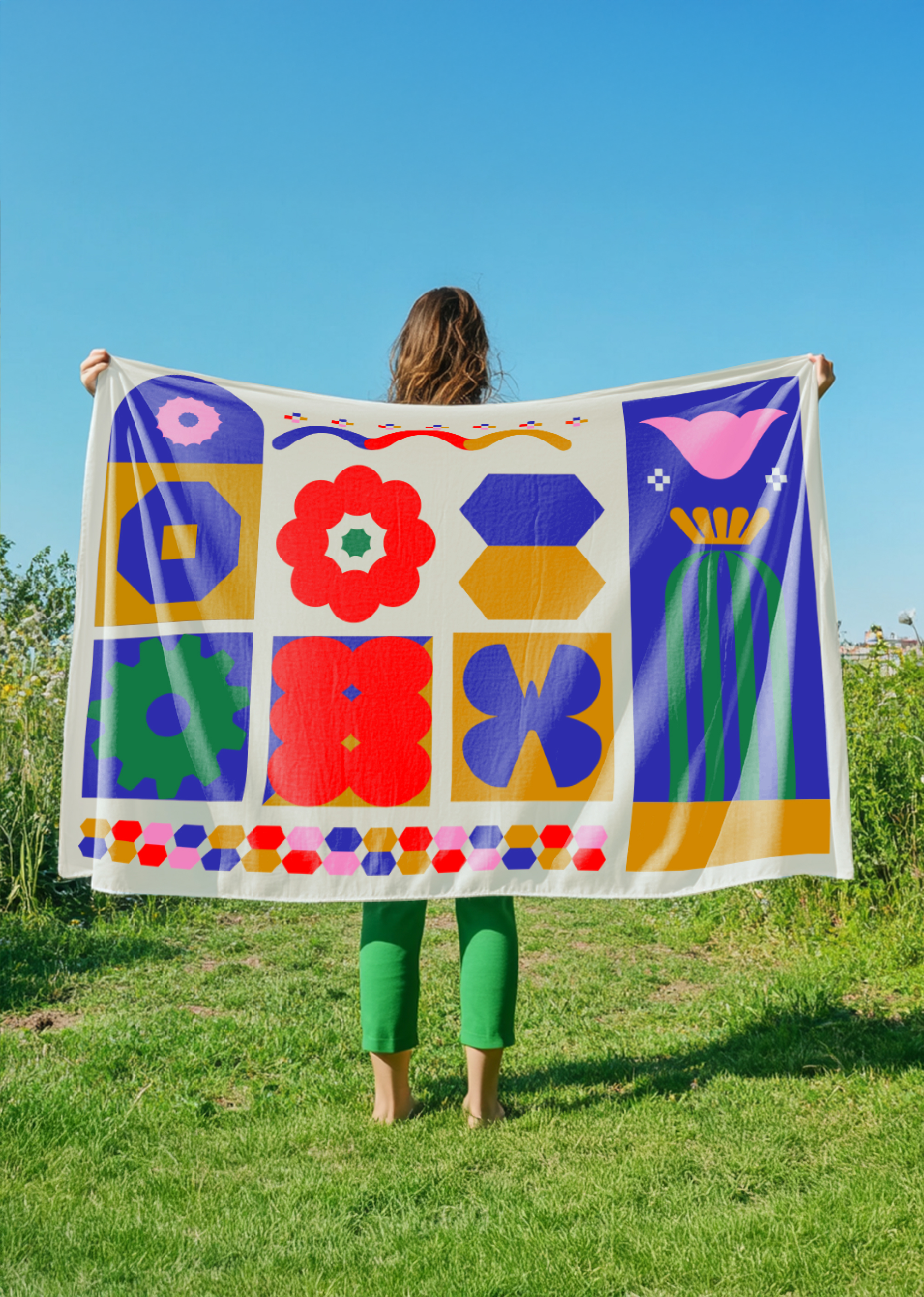 Persona de espaldas que sostiene una bandera con diferentes patrones y colores, en un campo con césped y cielo despejado.