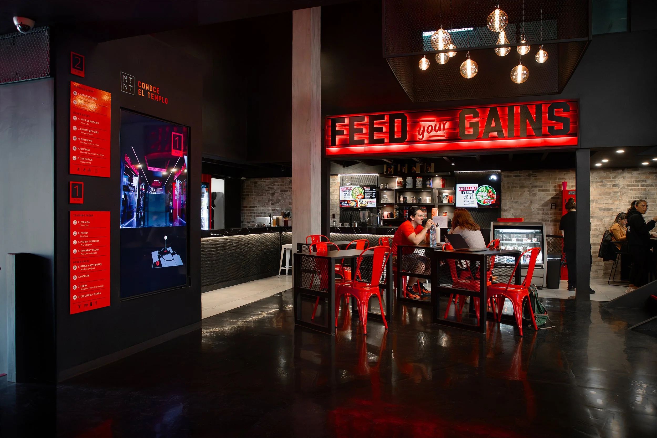 Interior de un gimnasio con un cartel luminoso que dice 'Feed your Gains', mesas, sillas rojas, monitores de pantalla y personas ejercitándose o socializando.