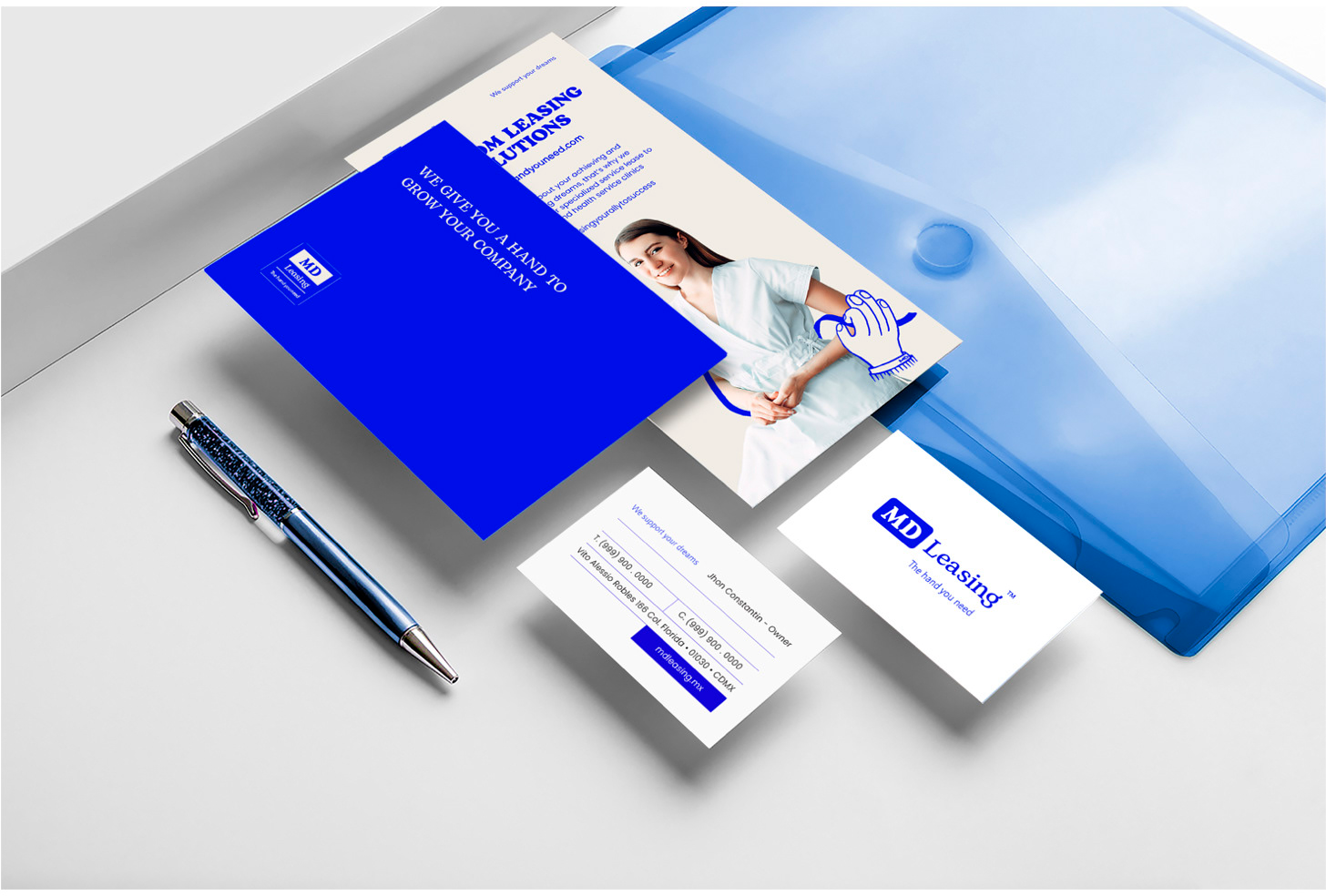 Material de oficina con folleto de MD Leasing, tarjeta de presentación y bolígrafo en una mesa blanca, y una carpeta azul translucida.