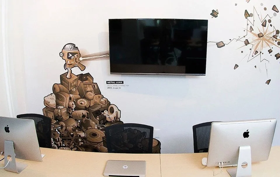 Sala de reuniones con dos computadoras Apple, una pared con mural de basura de metal formando una figura, y televisión montada.