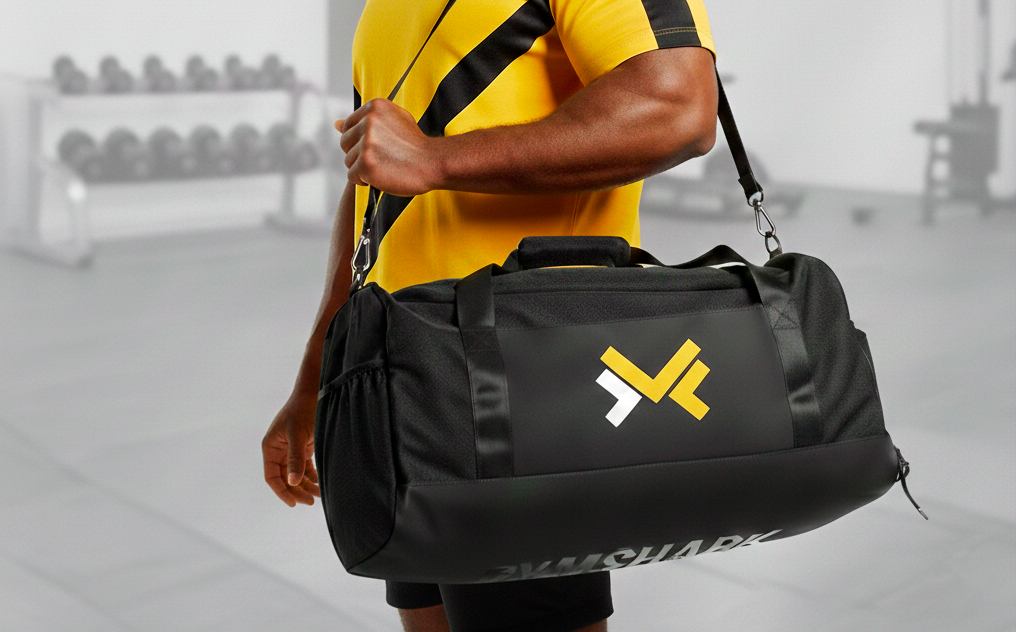 Persona con pantalones cortos y camiseta deportiva amarilla con rayas negras, sosteniendo una bolsa de deporte negra con un logotipo amarillo y blanco en un gimnasio.