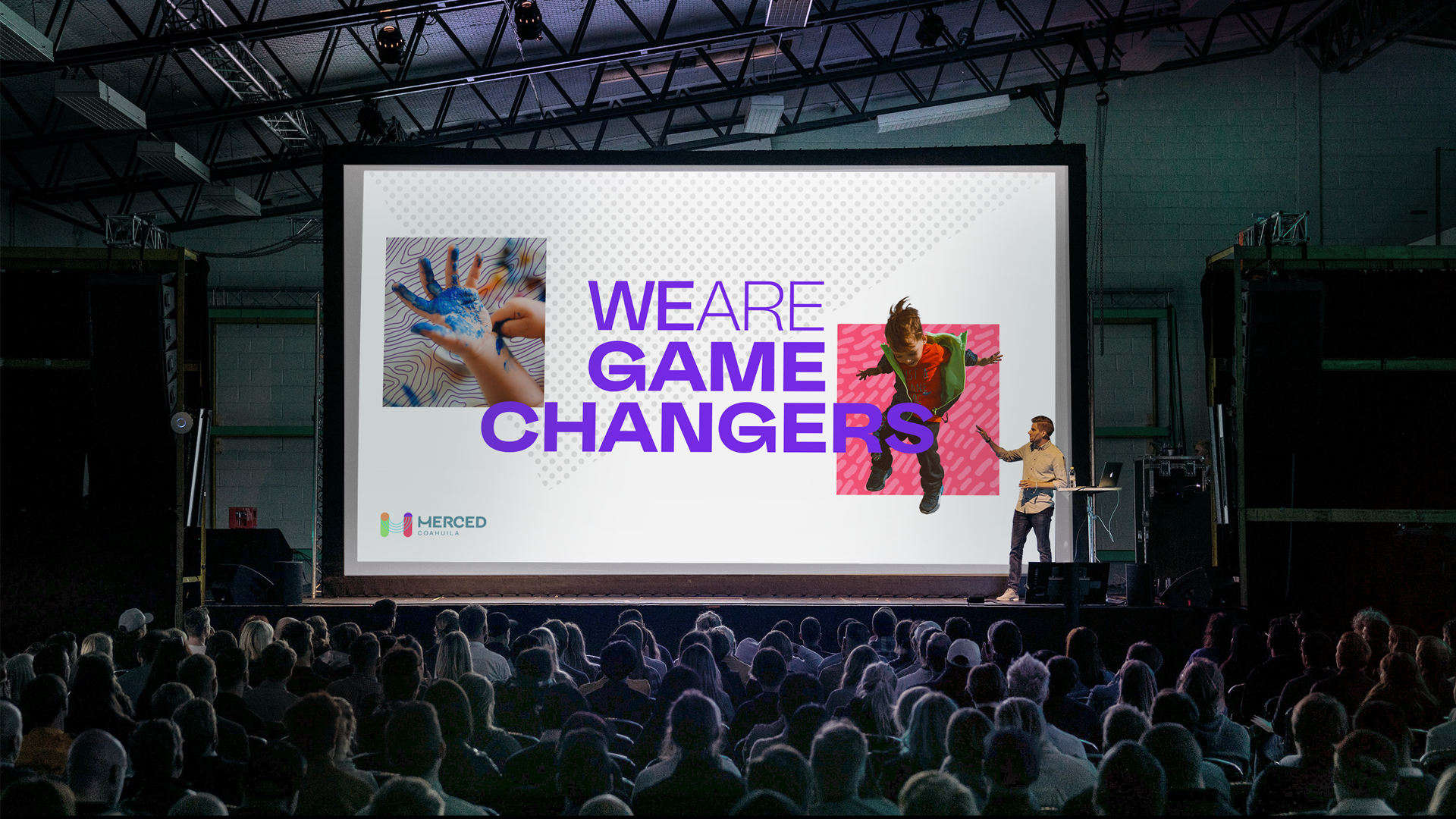 Presentación en un escenario con audiencia de muchas personas, fondo con pantalla grande que tiene el texto 'We are game changers' en letras moradas y dos imágenes de niños jugando, uno con pintura en las manos y otra saltando, en el logo de abajo que dice 'Merced Coahuila'.