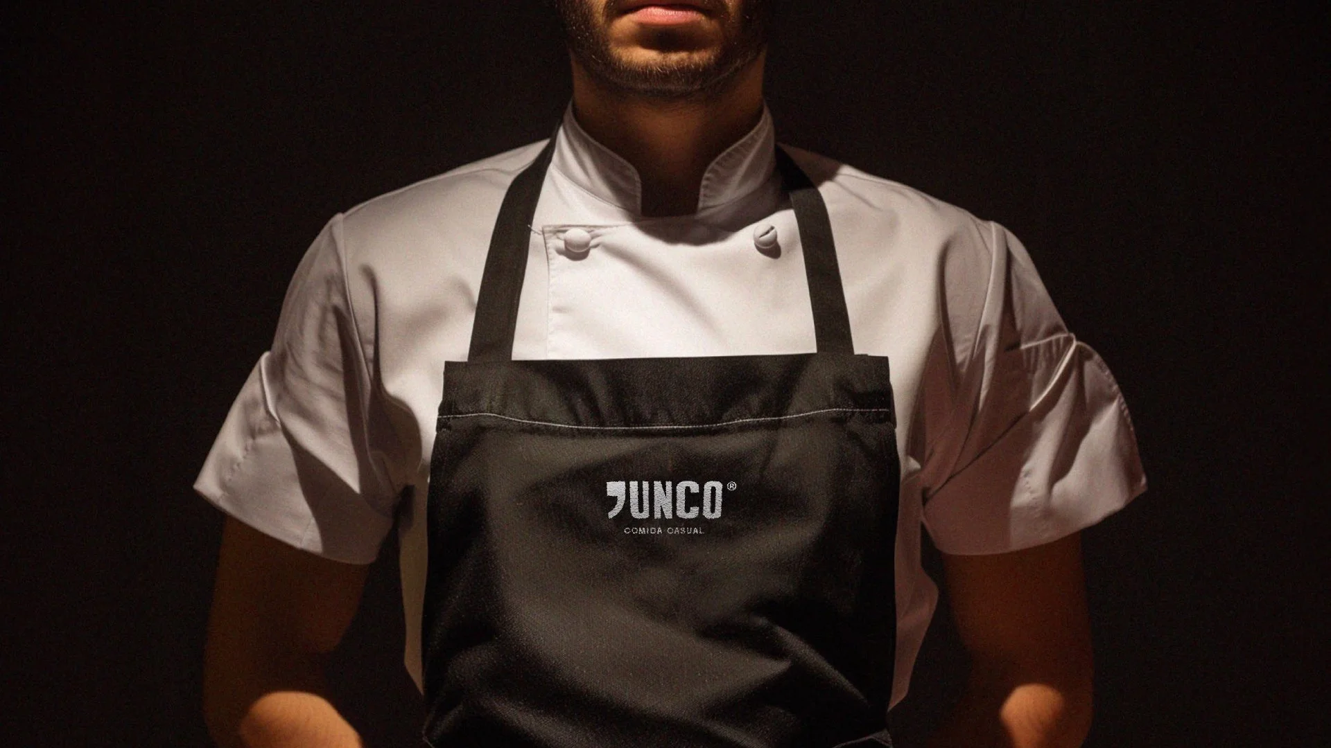 Persona con barba usando camiseta blanca y delantal negro que tiene el logo 'JUNCO' impreso en él, en un fondo oscuro.