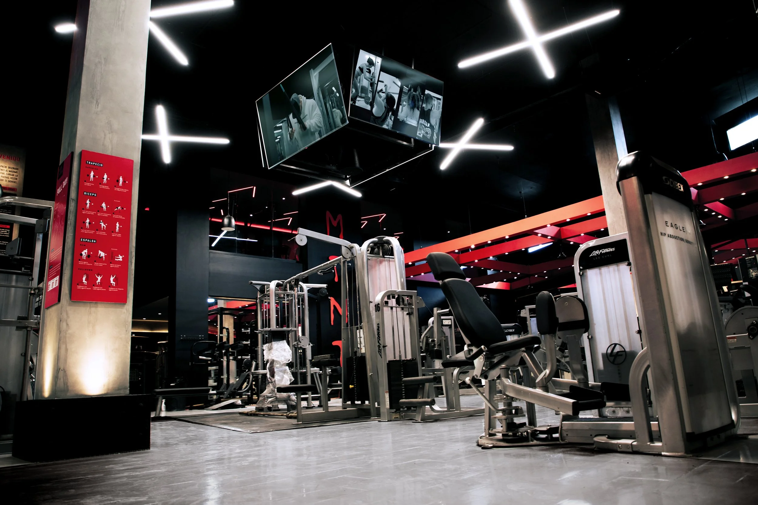 Área de gimnasio desierta con máquinas y equipo de ejercicio, paredes oscuras y luces modernas.
