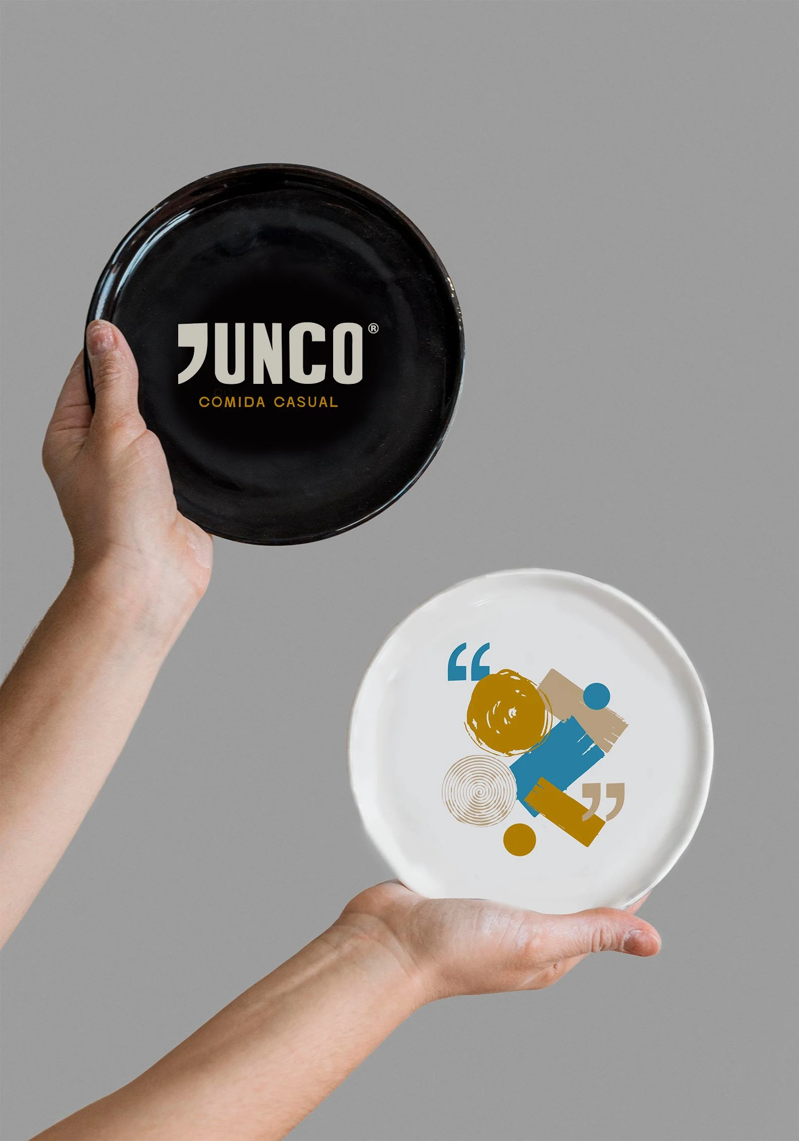 Manos sosteniendo platos, uno negro con el logo 'JUNCO Comida Casual' y otro blanco con diseño abstracto en colores azul, beige, y dorado.