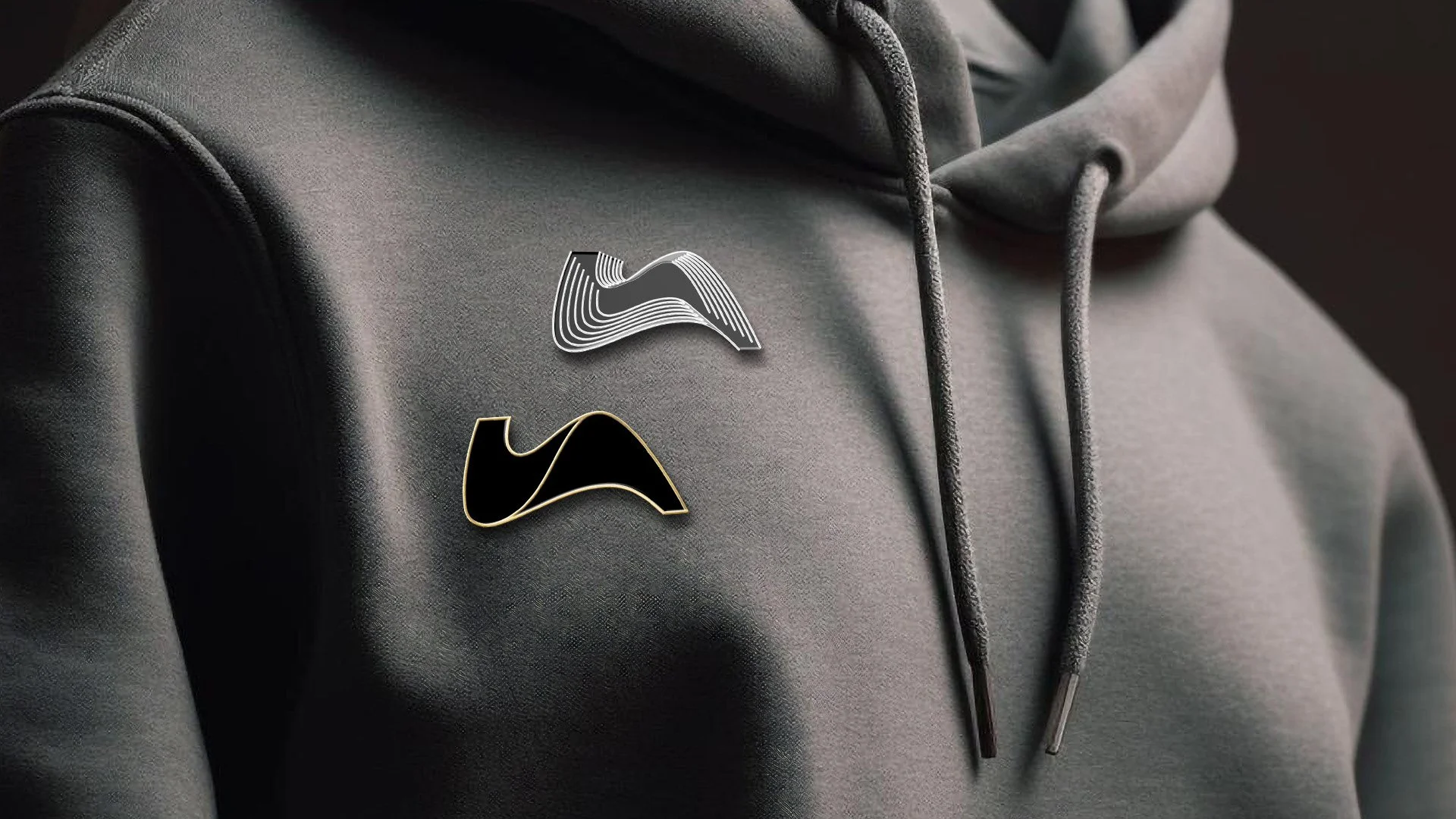 Dos pines con logotipos de Lopez Atilano en una chaqueta gris con capucha, uno en blanco y negro y otro en negro con borde dorado.