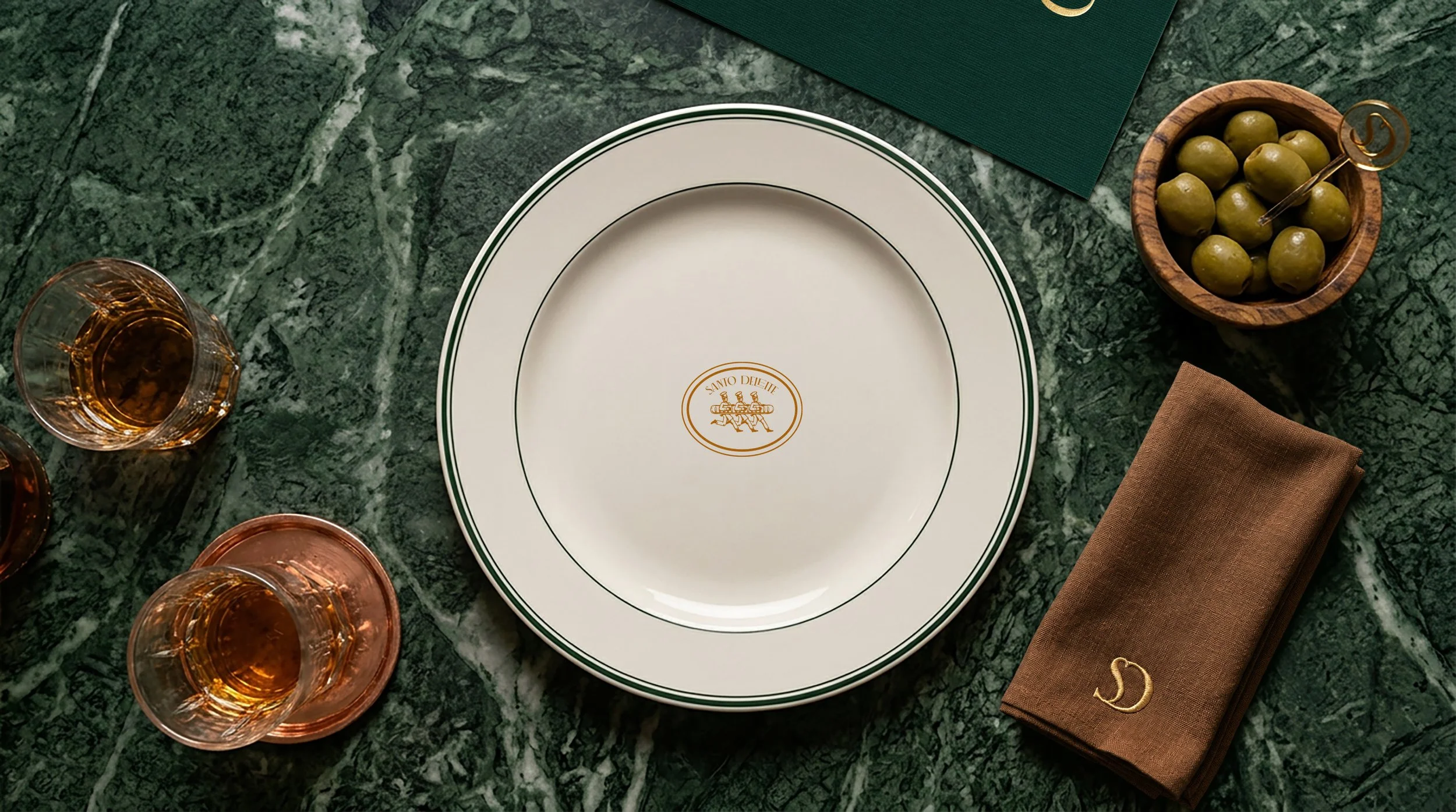 Lujosa mesa de comedor con plato blanco, vasos con bebida, aceitunas en un tazón de madera, cubiertos y servilleta de tela sobre mármol verde oscuro.