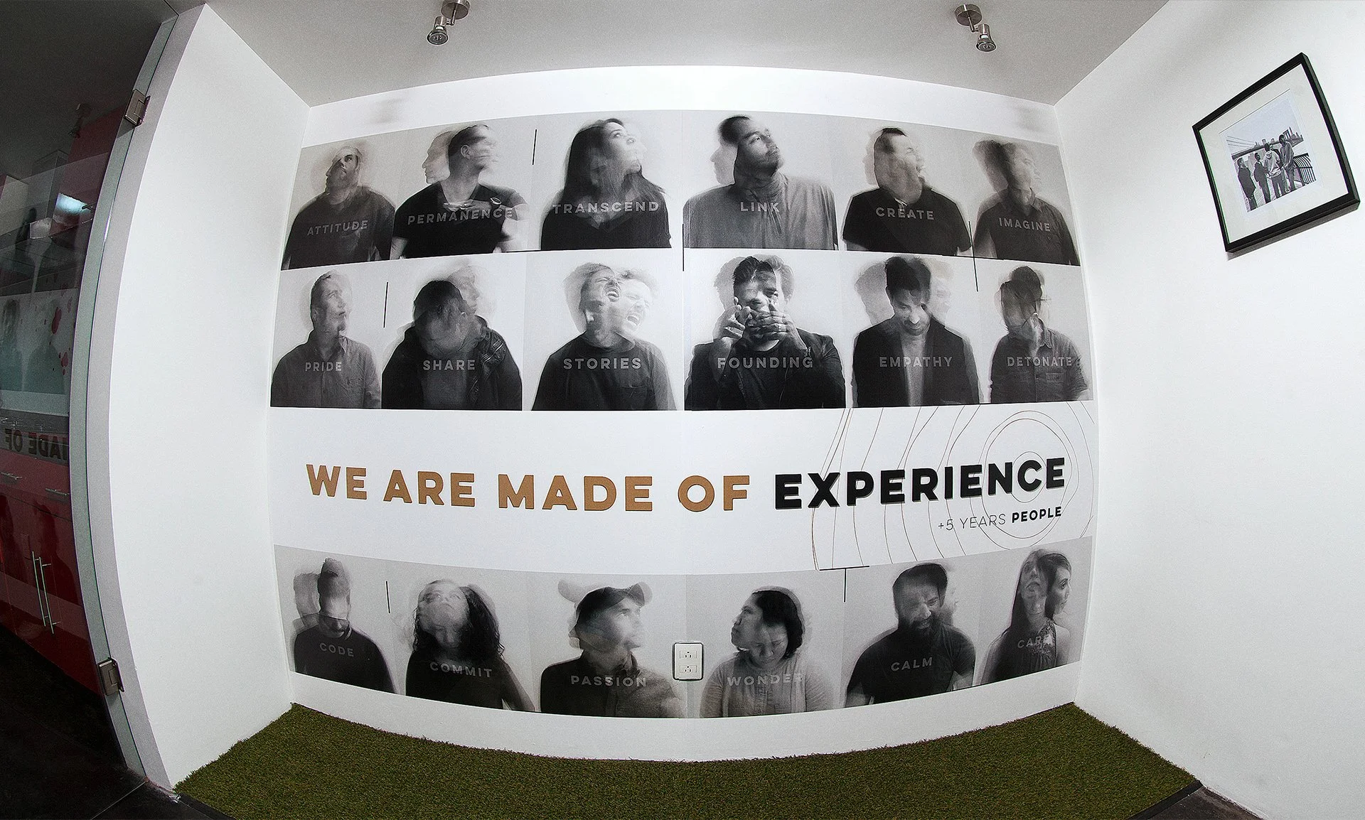Pared con retratos en blanco y negro de diferentes personas, cada una con palabras relacionadas con experiencias y cualidades como actitud, permanencia, trascender, vínculo, crear, imaginar, orgullo, compartir, historias, fundar, empatía, desactivar, código, compromiso, pasión, maravillar, calma. Texto en la parte inferior que dice "We are made of experience" y en español "Somos hechos de experiencia" con un mensaje adicional de +5 años de gente.