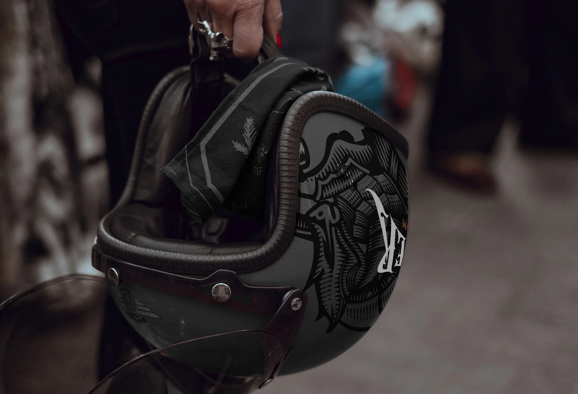 Casco de motocicleta con diseño en negro y gris y una ilustración de un caballero medieval en el costado, siendo sostenido por una mano con uñas pintadas de rojo, en un entorno con fondo desenfocado.
