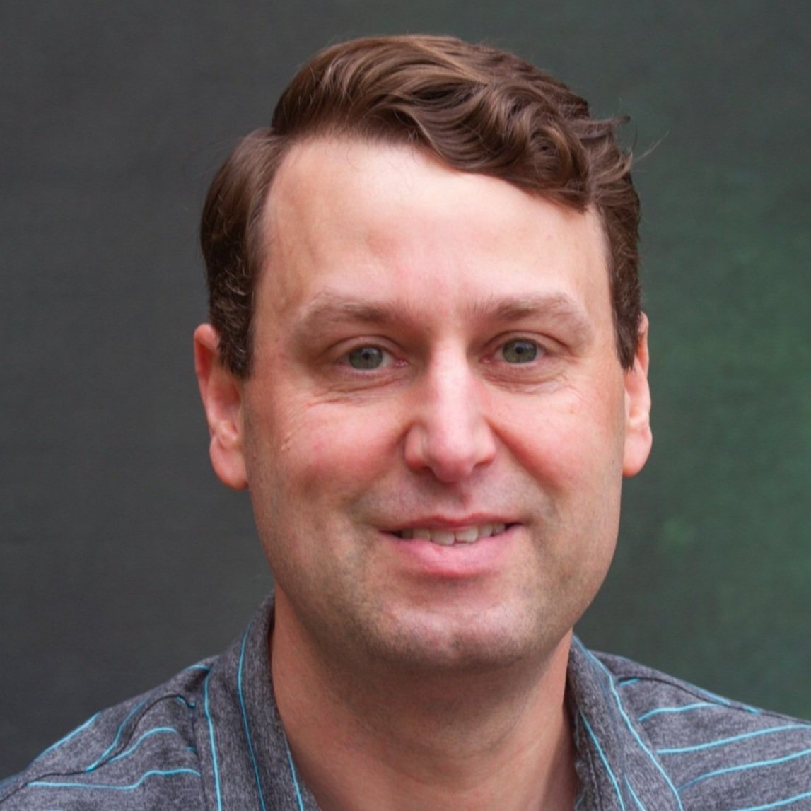 Close-up portrait of a smiling man with light skin, brown hair, and green eyes, wearing a gray shirt with blue stripes, against a blurred dark green background.