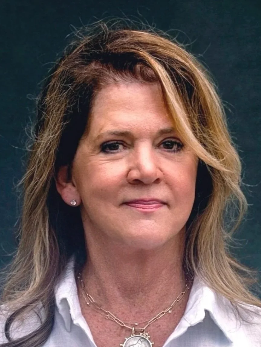 A woman with shoulder-length brown hair and light skin, wearing a white collared shirt, a necklace with a gear-shaped pendant, and earrings, standing against a dark background.
