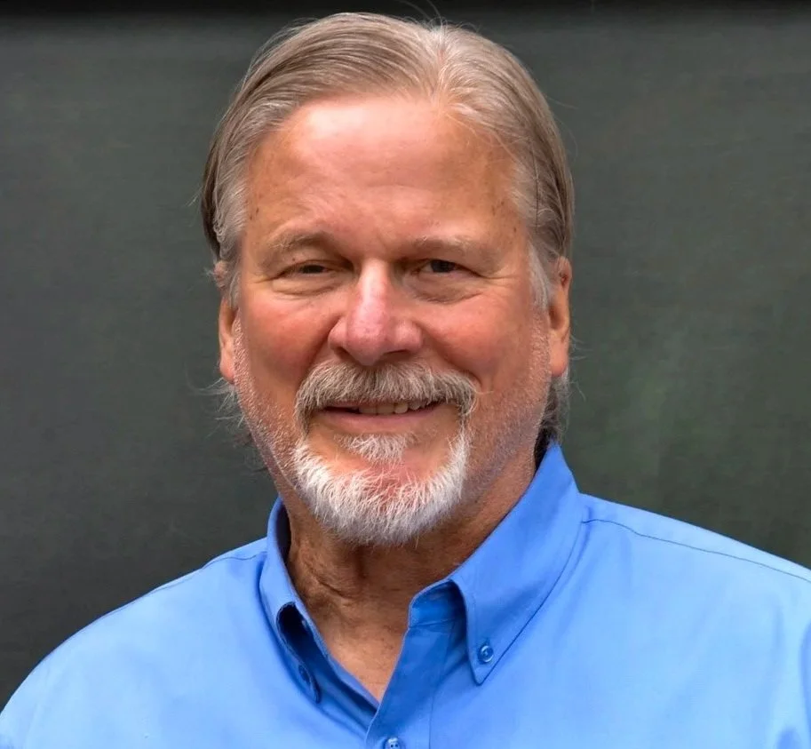 A smiling man with gray hair and beard, wearing a blue button-up shirt, standing against a dark background.