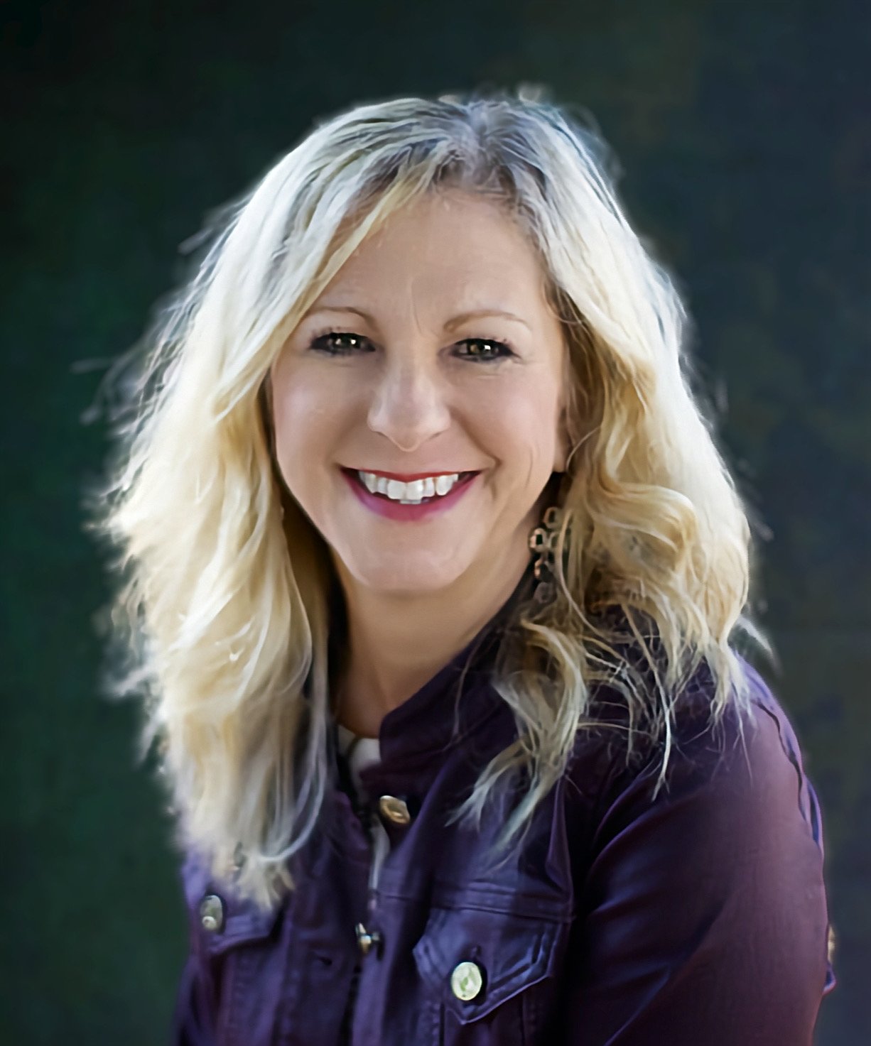A woman with blonde, shoulder-length curly hair, smiling in front of a dark background.