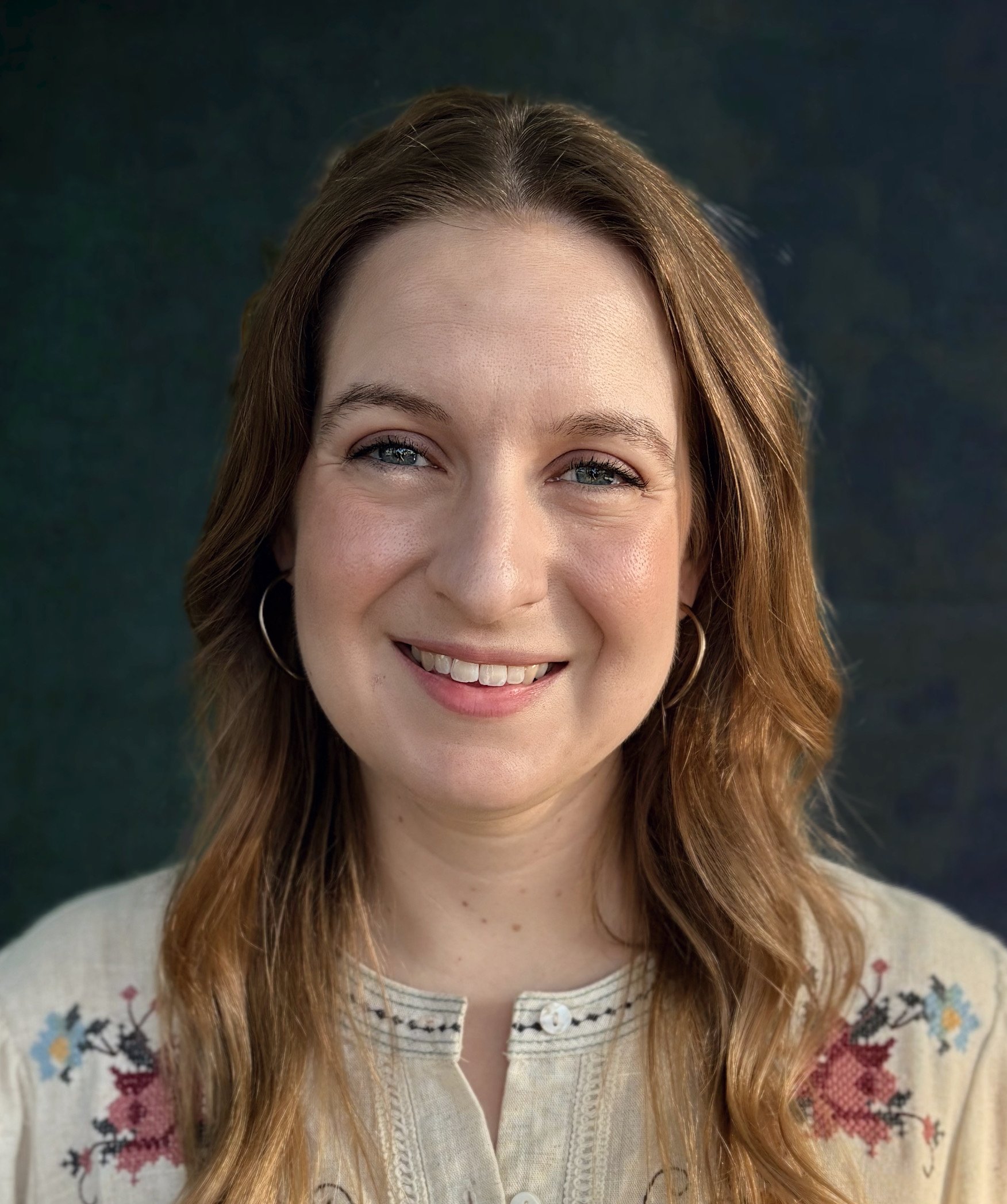 A woman with long light brown hair, smiling, wearing purple clothing and a pearl earring, against a dark, blurry background.